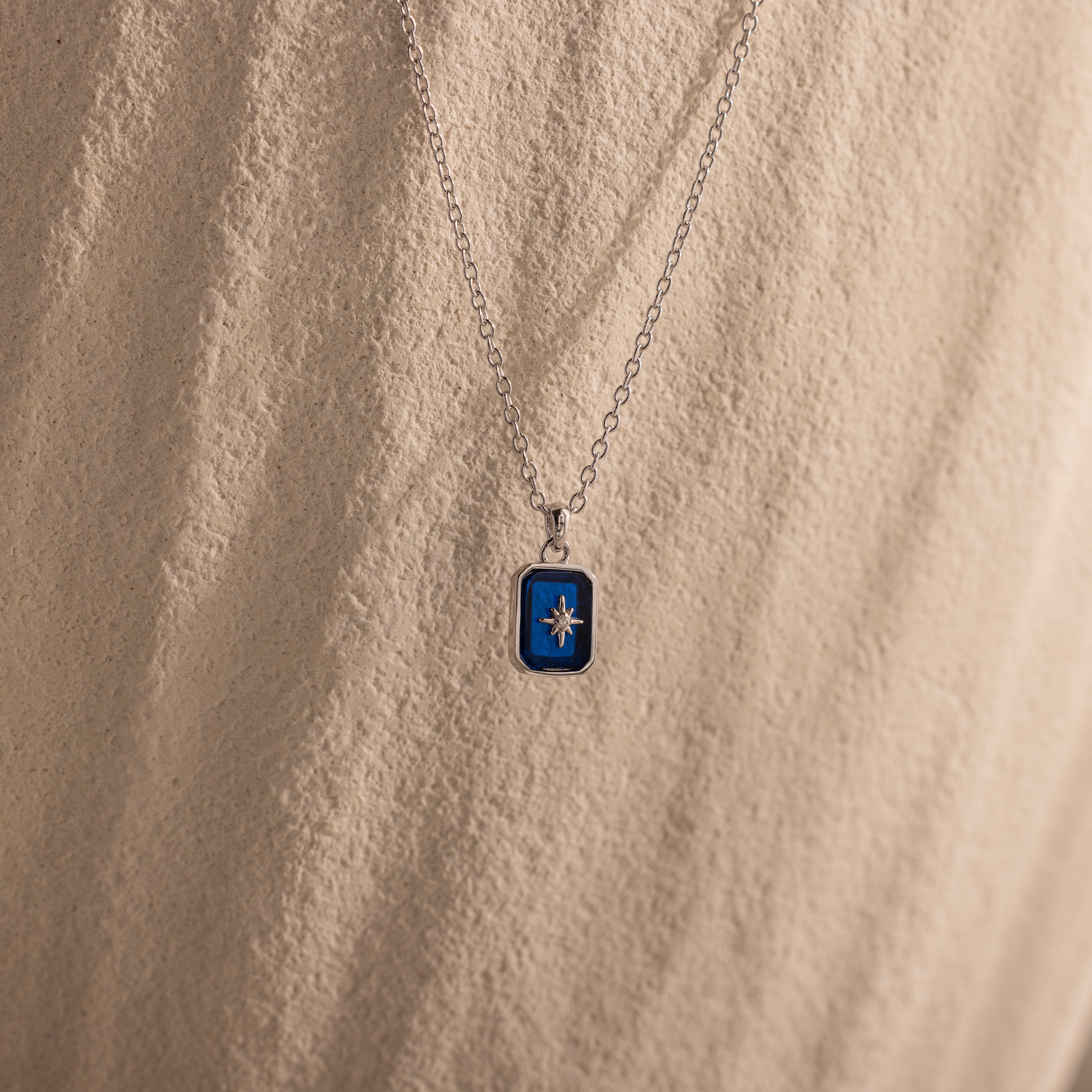 The North Star Sapphire Necklace features a blue rectangular crystal pendant with a diamond-set North Star, displayed on a textured beige surface.