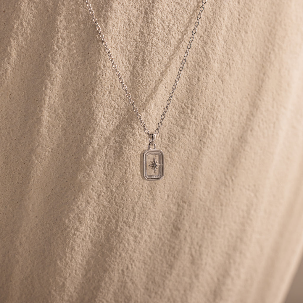 The North Star Diamond Necklace features a rectangular crystal pendant with a delicate diamond-set North Star design, showcased against a textured beige background.