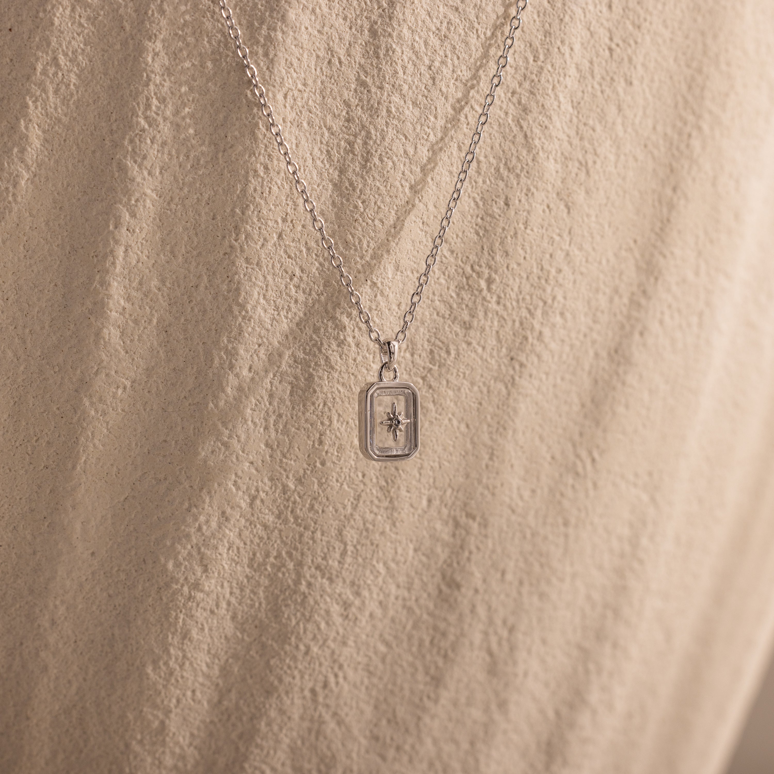 The North Star Diamond Necklace features a rectangular crystal pendant with a delicate diamond-set North Star design, showcased against a textured beige background.