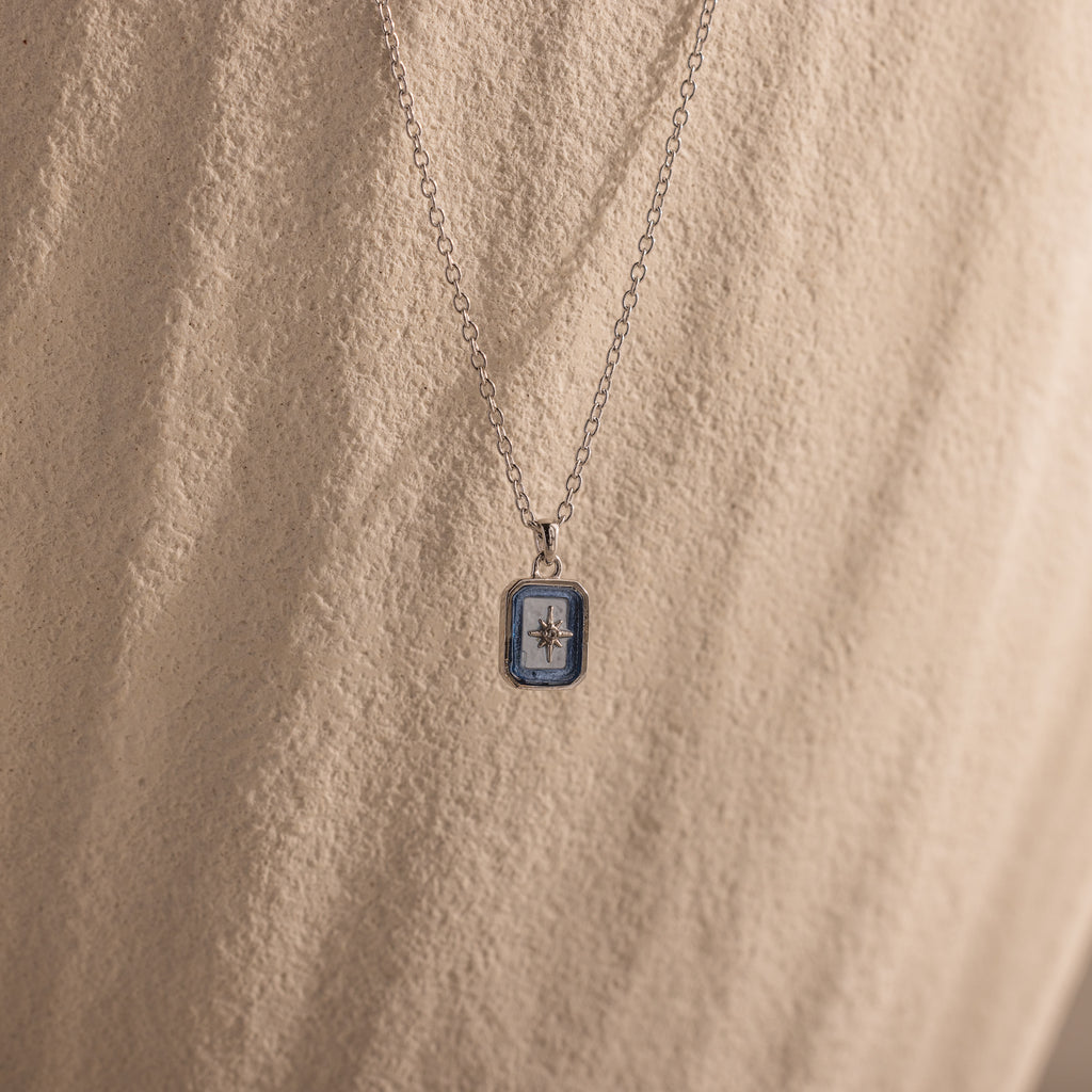North Star Aquamarine Necklace with a small rectangular pendant and a North Star design, accented by aquamarine stones, set against a textured beige background.