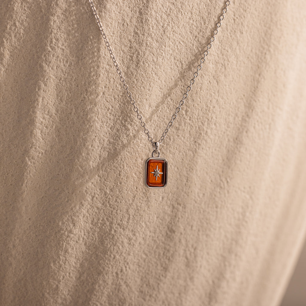 The North Star Garnet Necklace features a silver chain with a rectangular orange crystal pendant and a small star, displayed on a textured beige surface.