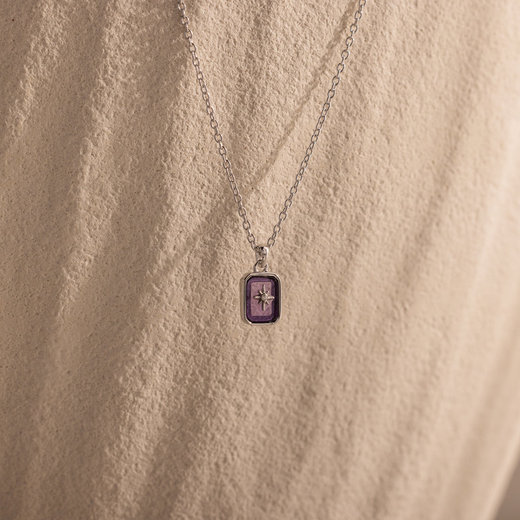 The North Star Amethyst Necklace, featuring a purple rectangular crystal pendant, is displayed on a textured beige background.