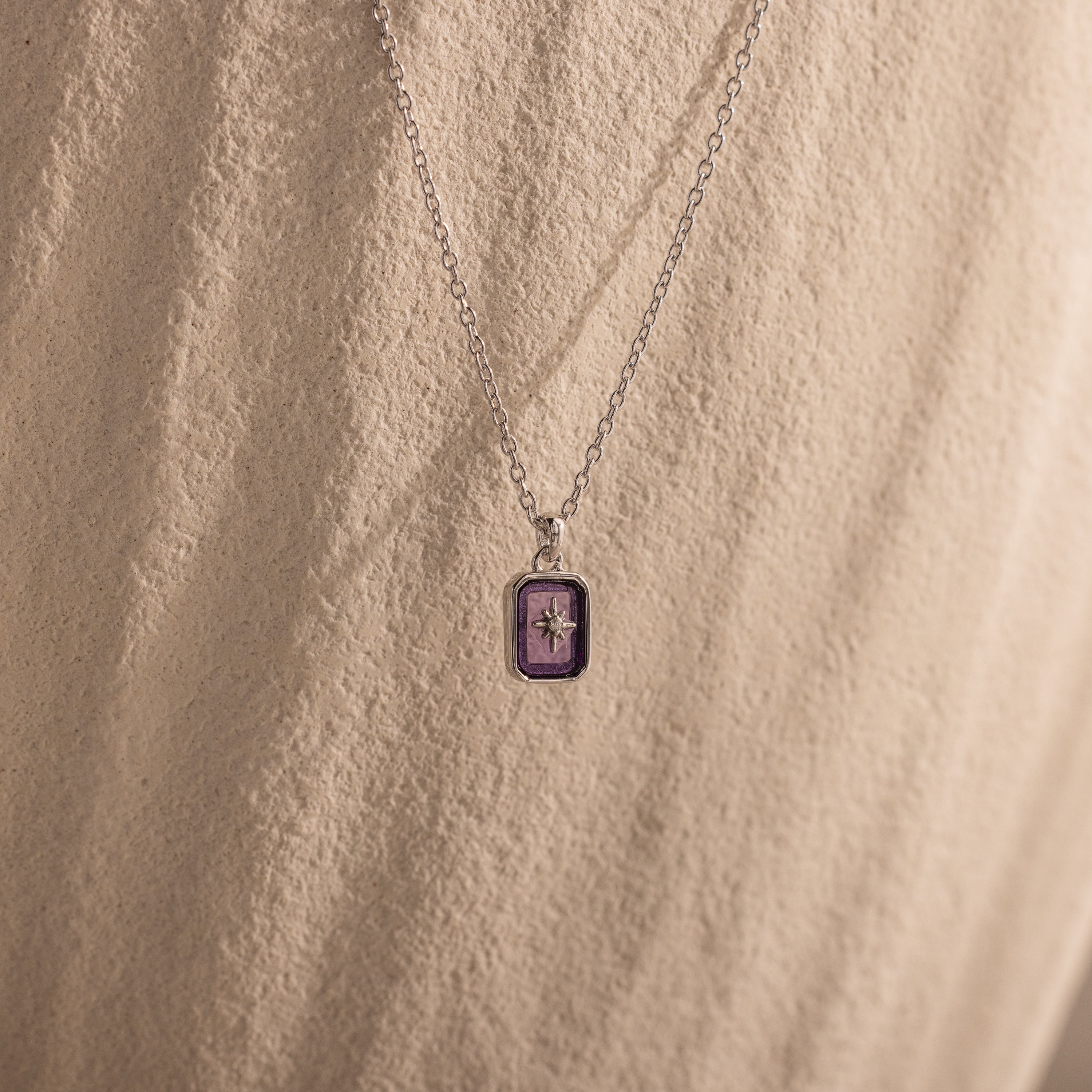 The North Star Amethyst Necklace, featuring a purple rectangular crystal pendant, is displayed on a textured beige background.