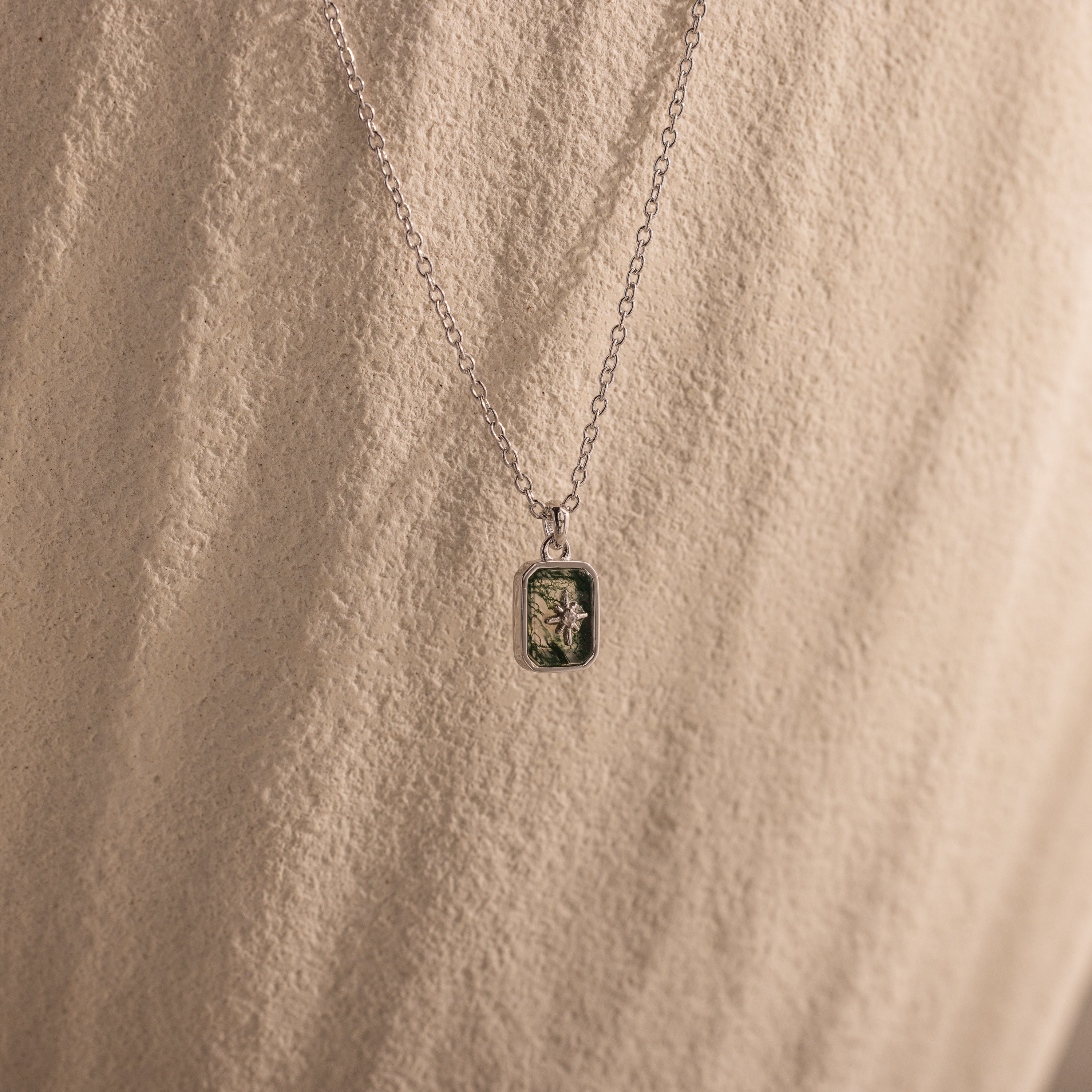 The North Star Gemstone Necklace features a rectangular crystal pendant with a small plant, displayed against a neutral textured background.