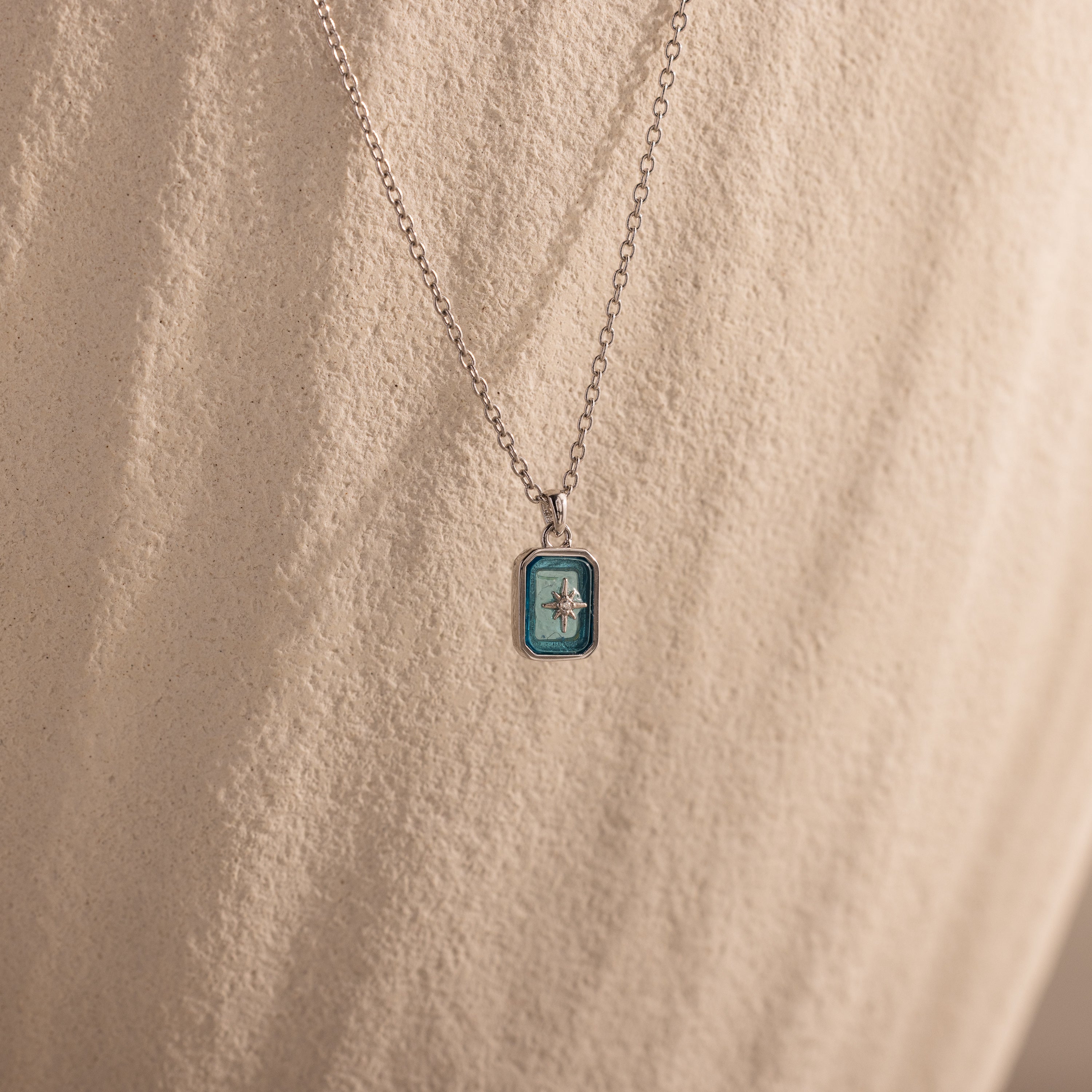 The North Star Blue Zircon Necklace features a small blue rectangular crystal pendant with a star design, shown against a textured beige background.