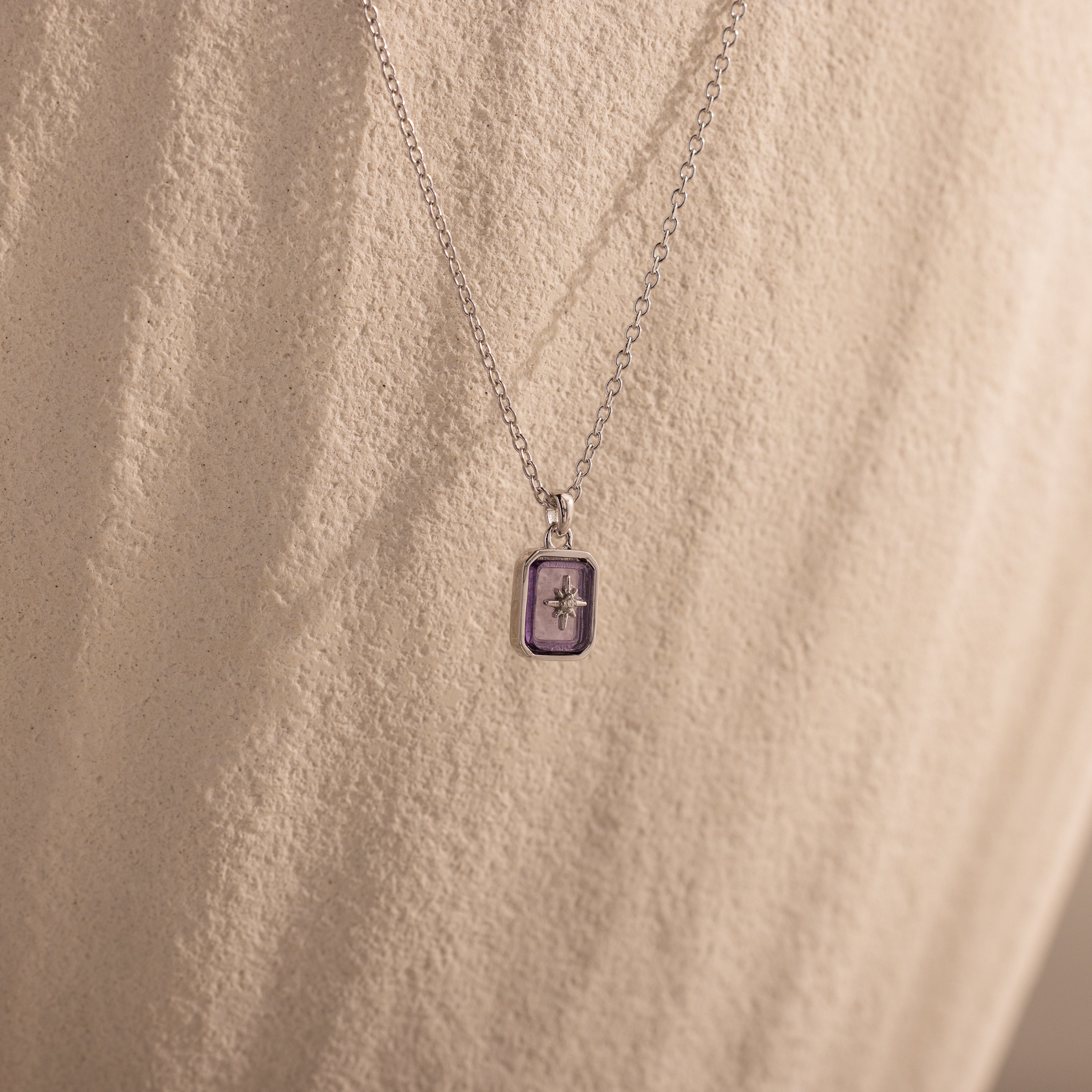 The North Star Alexandrite Necklace features a silver chain with a purple rectangular crystal pendant and a small star detail, displayed on a textured beige surface.