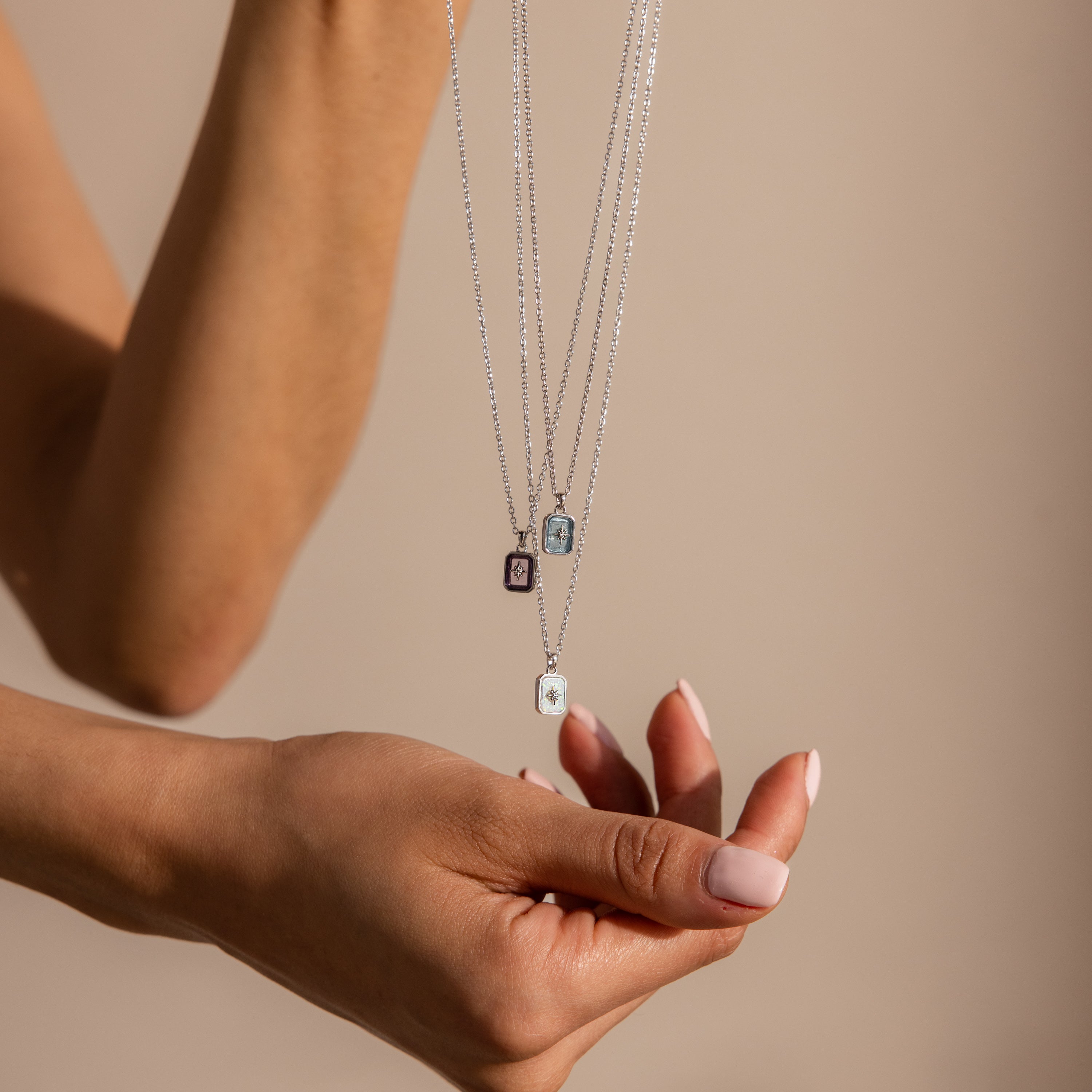A hand holds three silver chain necklaces, each featuring a North Star Opal Necklace with a rectangular crystal pendant, against a beige background.