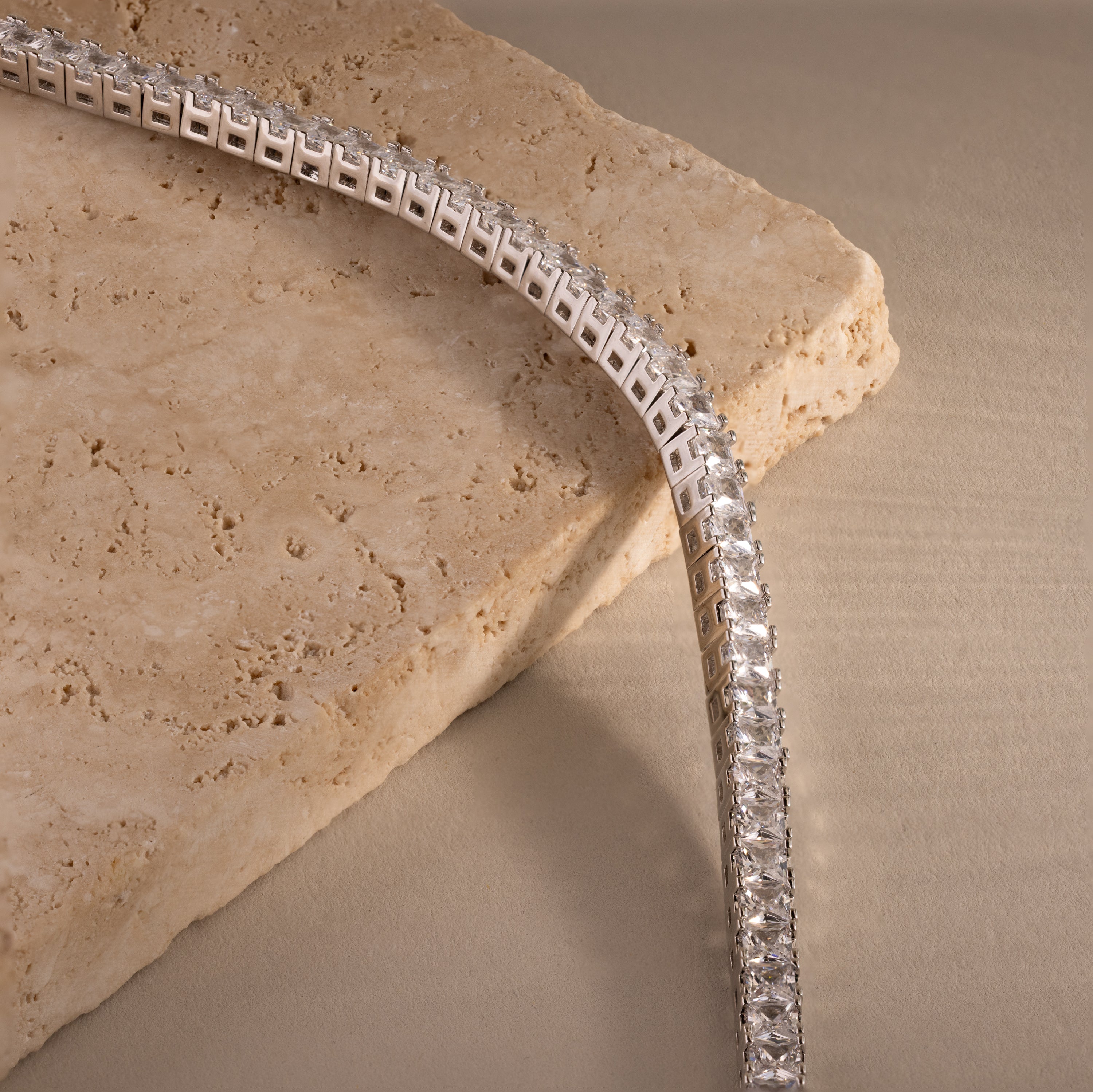 The Princess Diamond Tennis Bracelet in White Gold, featuring square-cut stones, is displayed on a textured beige stone surface.