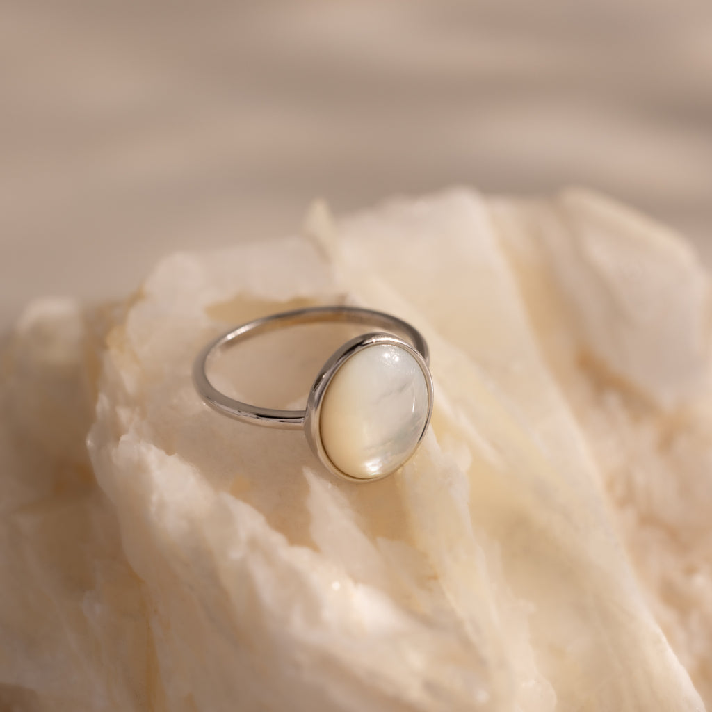 The Mother of Pearl Signet Ring in Sterling Silver, featuring a vintage-inspired round milky gemstone, is showcased on a creamy textured stone surface.