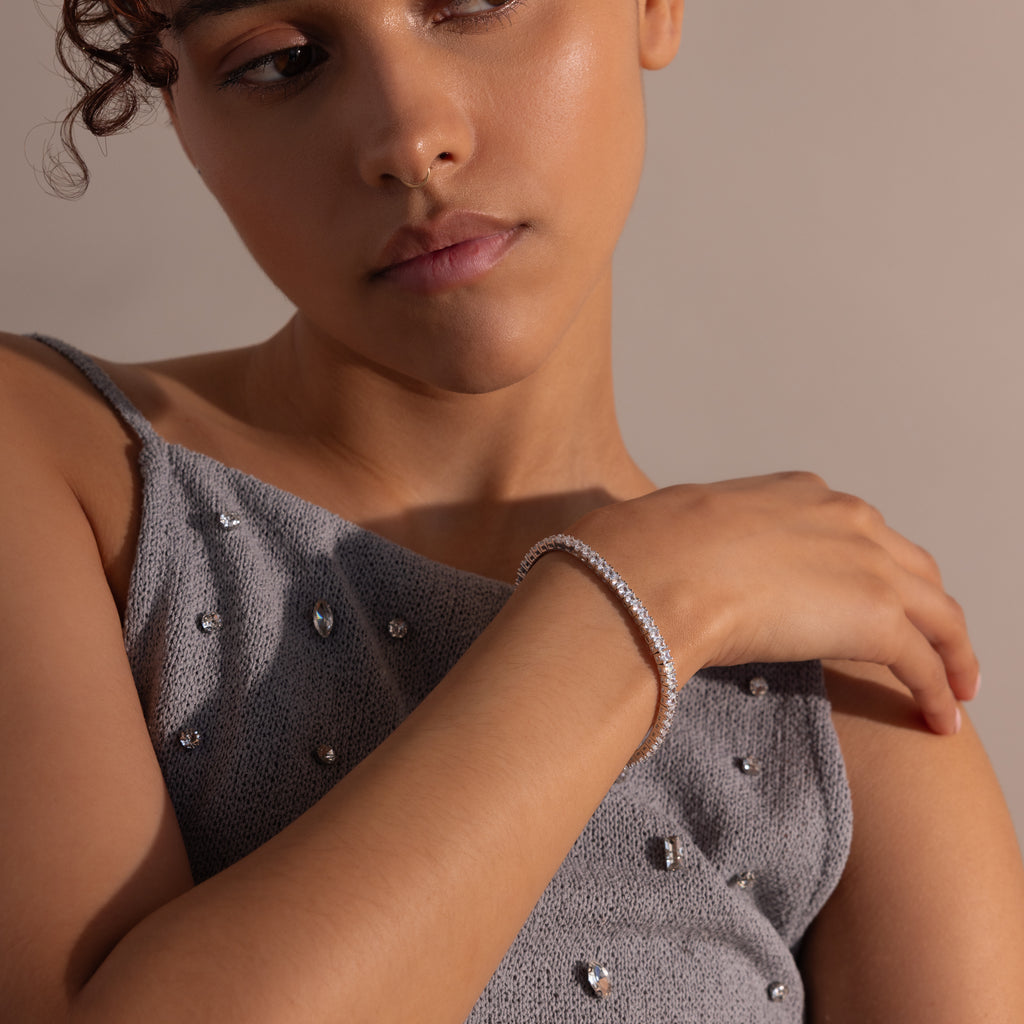 A woman in a gray beaded top wears the Princess Diamond Tennis Bracelet in White Gold, resting her hand on her shoulder against a neutral background.