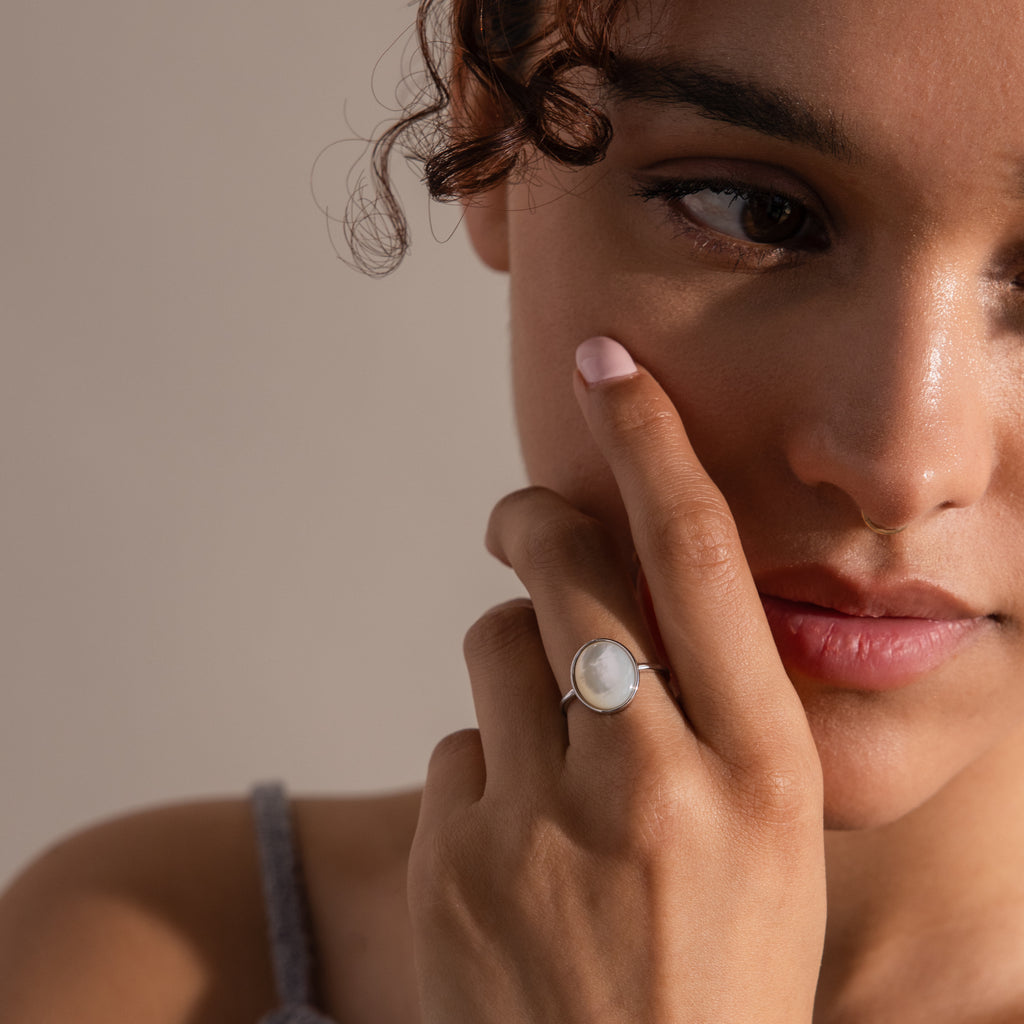 A woman with curly hair gently touches her face, wearing the vintage-inspired Mother of Pearl Signet Ring in Sterling Silver, featuring a striking white stone under soft lighting.