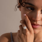 A woman with curly hair gently touches her face, wearing the vintage-inspired Mother of Pearl Signet Ring in Sterling Silver, featuring a striking white stone under soft lighting.