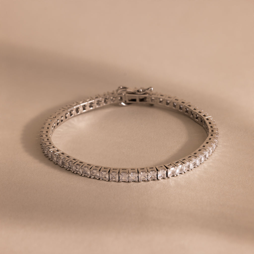 The Princess Diamond Tennis Bracelet, featuring sparkling princess-cut diamonds, is showcased on a beige background.