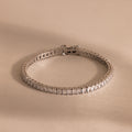 The Princess Diamond Tennis Bracelet, featuring sparkling princess-cut diamonds, is showcased on a beige background.