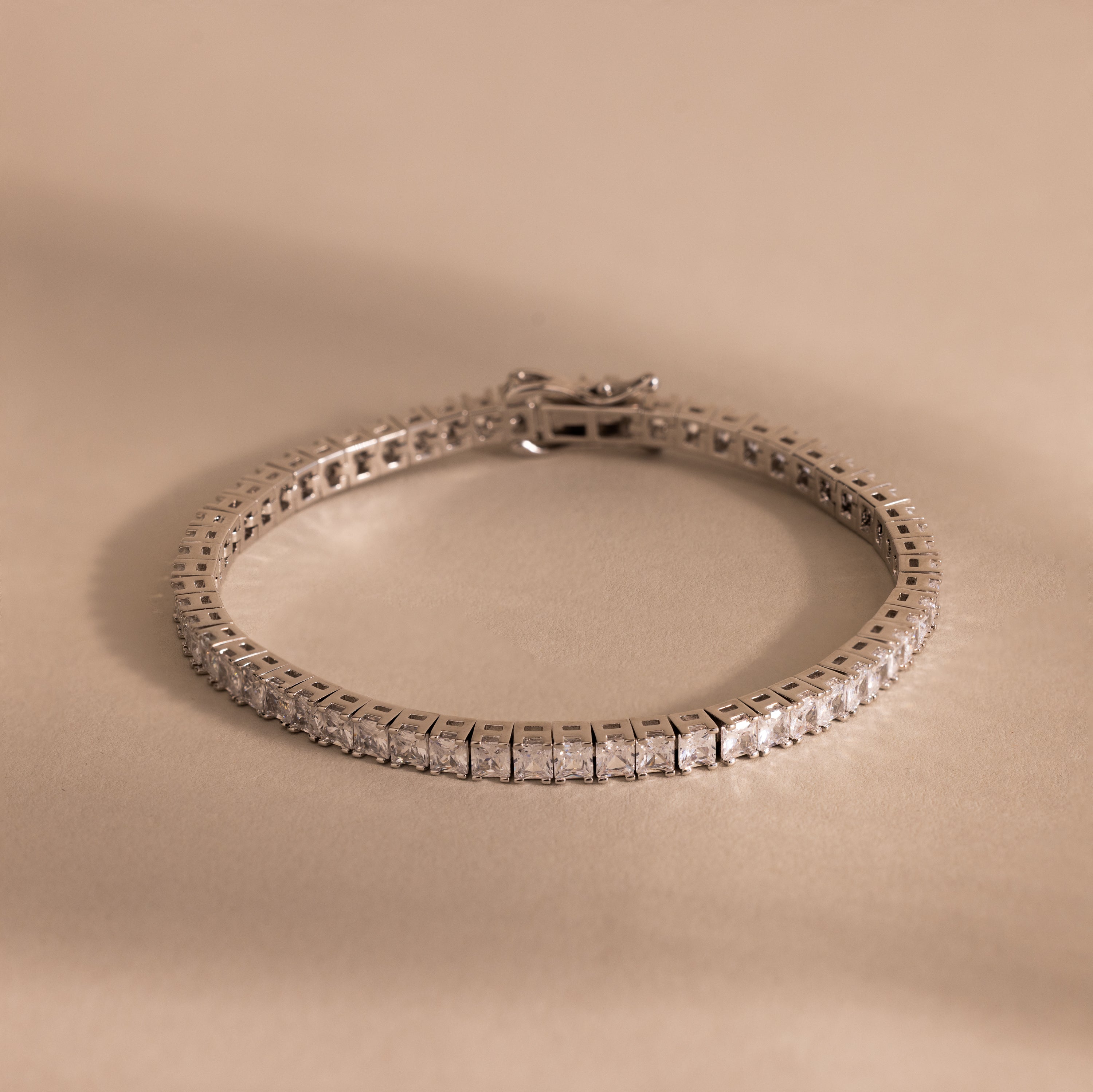 The Princess Diamond Tennis Bracelet, featuring sparkling princess-cut diamonds, is showcased on a beige background.
