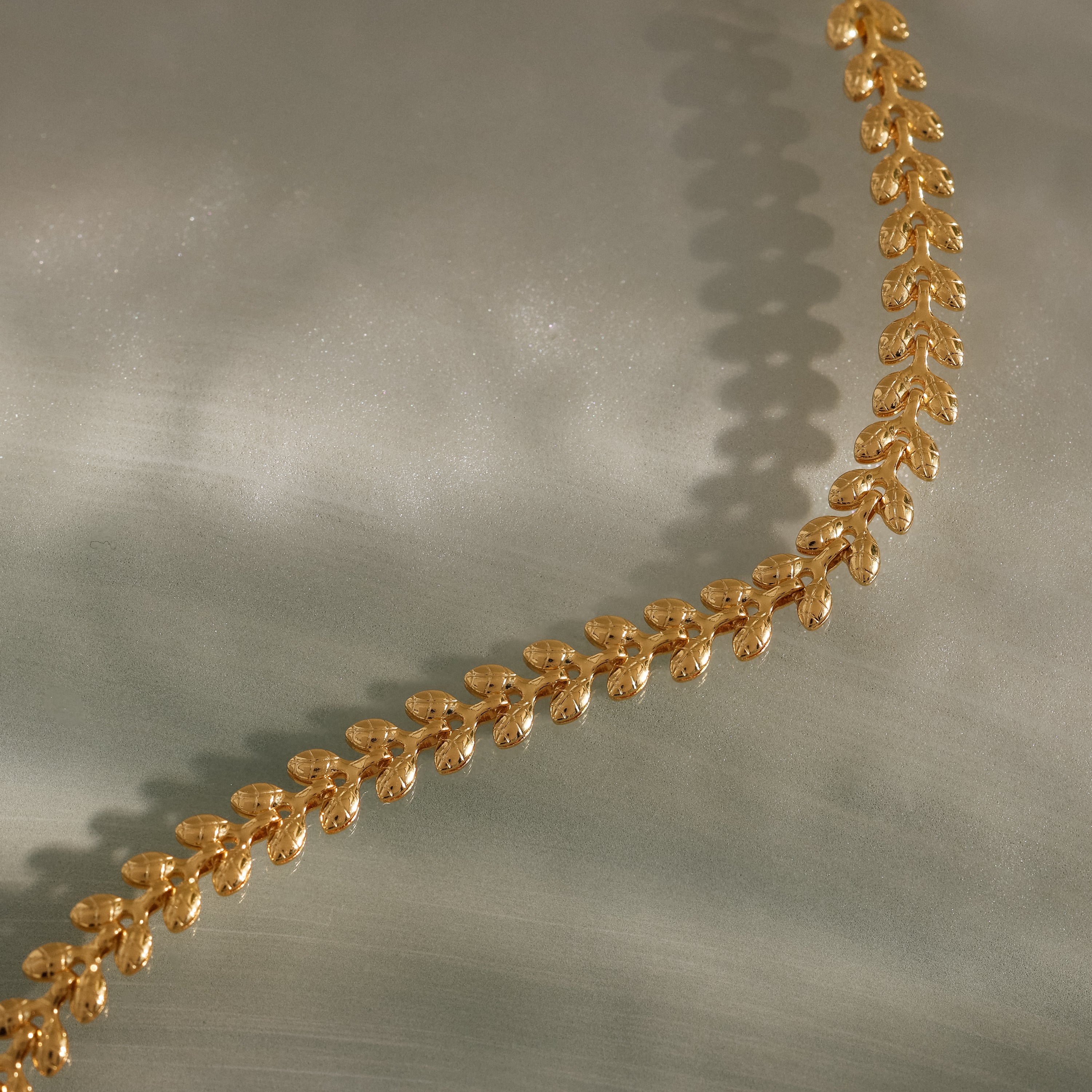 The Fishtail Chain Bracelet, featuring detailed craftsmanship and a leaf-shaped link design, rests on a soft, light green textured surface.