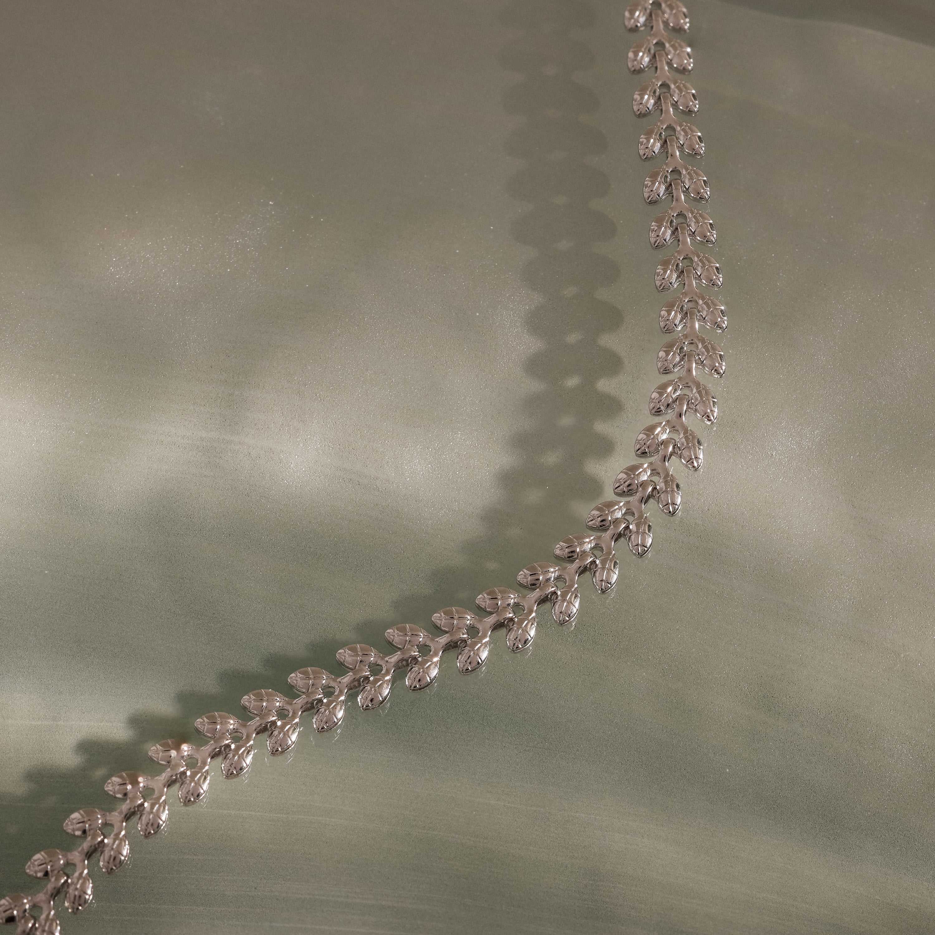 The Fishtail Chain Bracelet in Silver features interwoven links and a delicate leaf pattern, elegantly displayed on soft pale green fabric—an ideal gift for any occasion.
