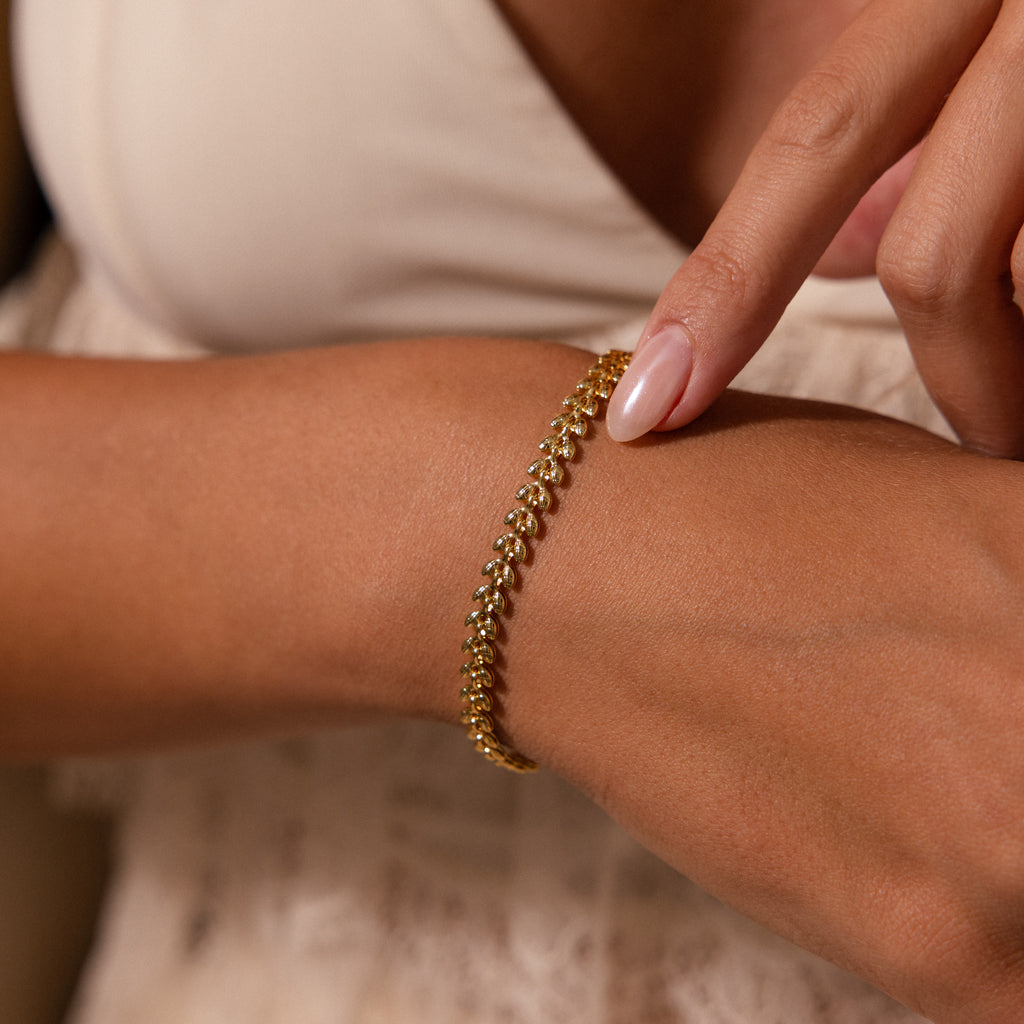 A person in a light, sleeveless top touches their gold Fishtail Chain Bracelet, highlighting its cascading texture and detailed craftsmanship.