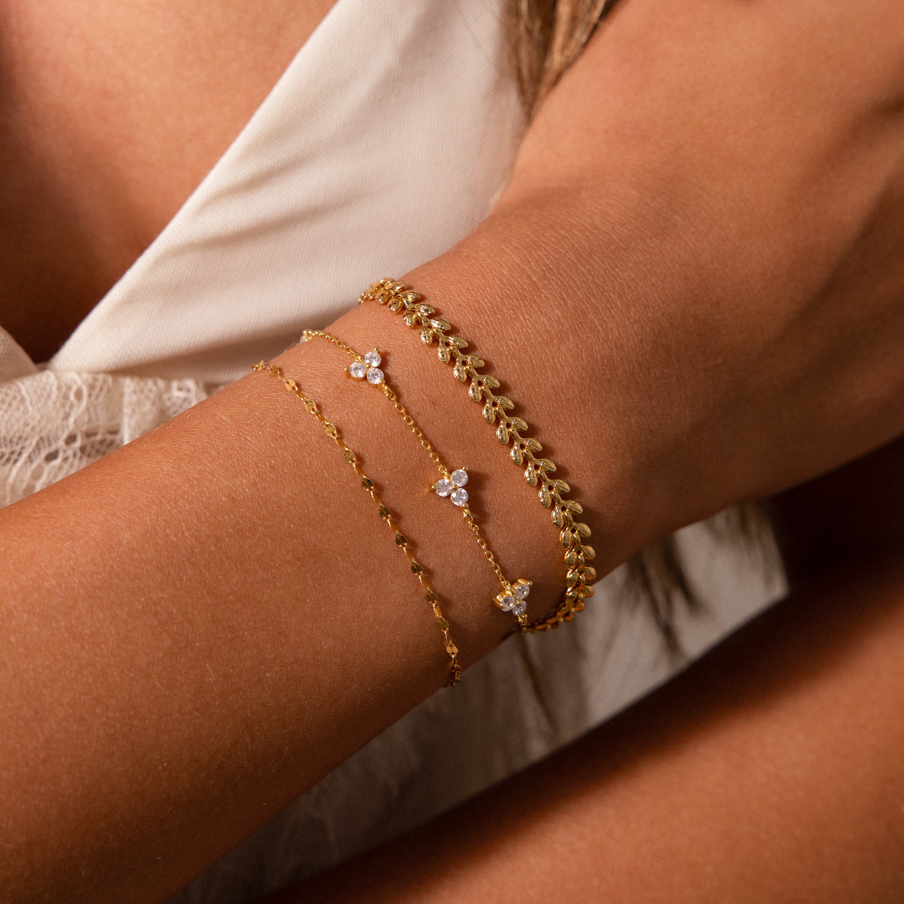 A woman in a white lace top wears three delicate gold bracelets, including the Fishtail Chain Bracelet featuring cascading texture and intricate charms on her wrist.