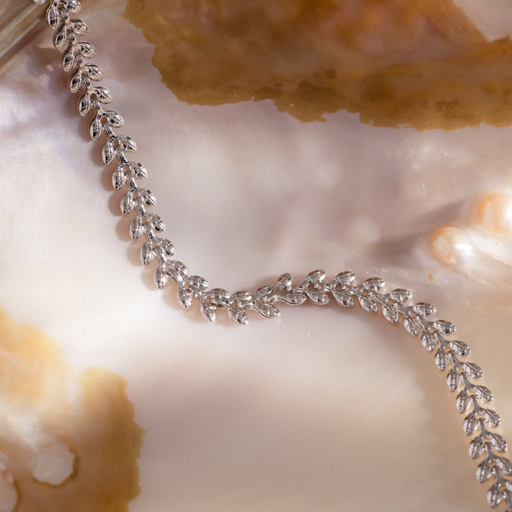 The Fishtail Chain Bracelet in Silver, featuring interwoven links, is displayed on a shiny iridescent shell with beige accents beside a silver leaf-patterned necklace.