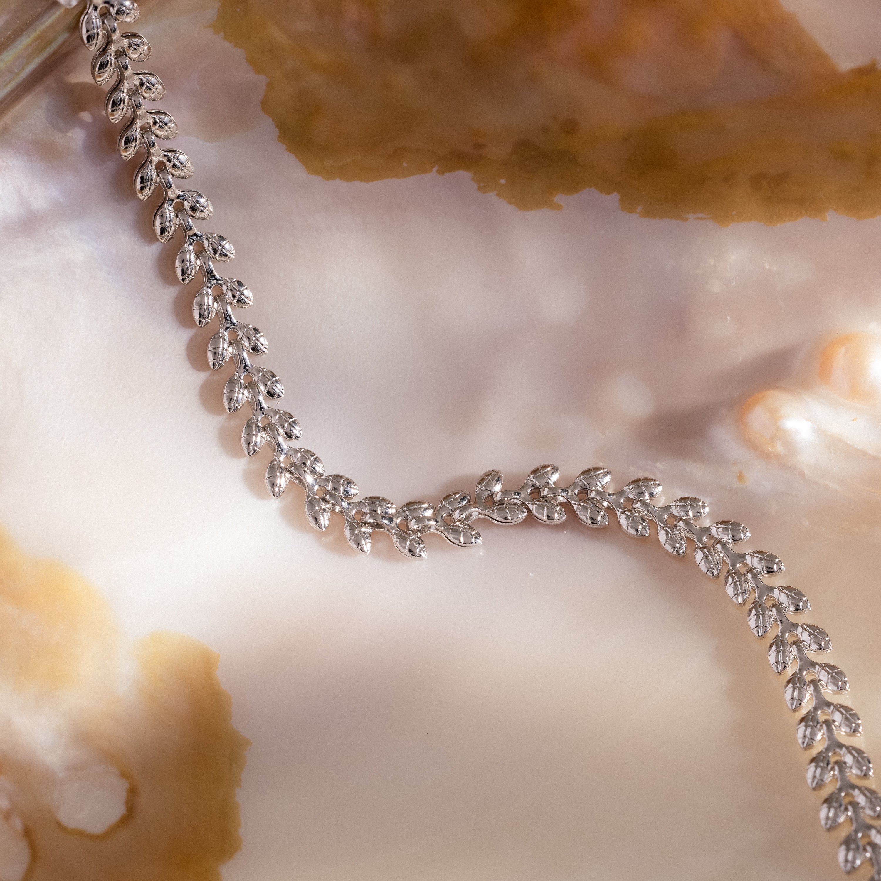 The Fishtail Chain Bracelet in Silver, featuring interwoven links, is displayed on a shiny iridescent shell with beige accents beside a silver leaf-patterned necklace.