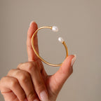 A hand holds the Serenity Pearl Cuff Bracelet, a gold open band with pearls on each end, highlighting modern pearl jewelry against a neutral background.