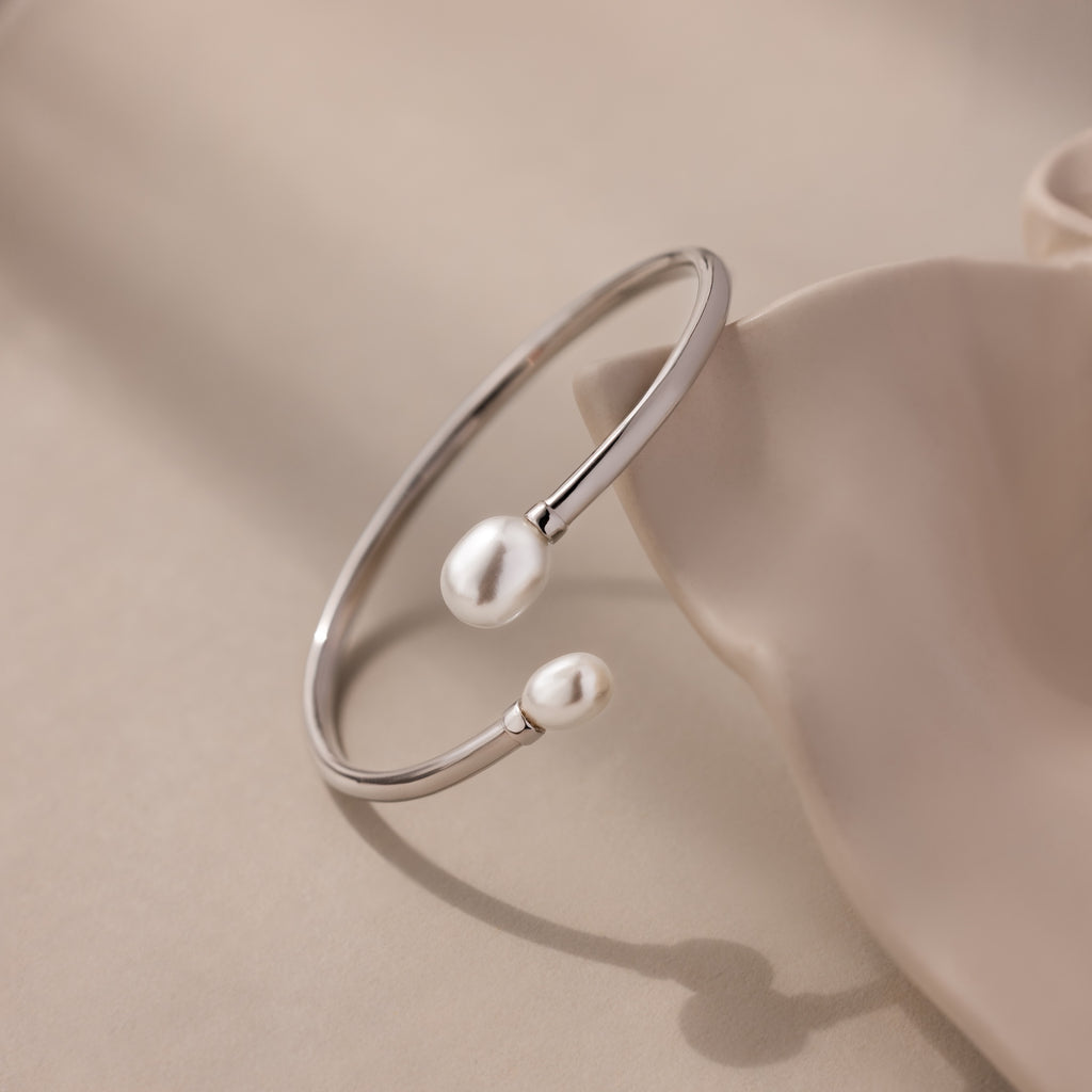 The Serenity Pearl Cuff Bracelet in Silver features an open band with two luminous pearls, elegantly displayed on a soft beige surface.