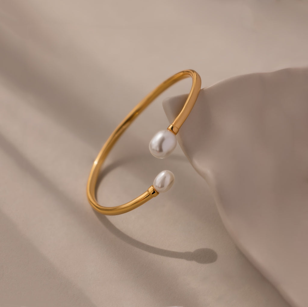 The Serenity Pearl Cuff Bracelet in 18K Gold features an open band with two white pearls on each end, elegantly displayed on a neutral beige surface—a modern statement piece.