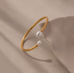 The Serenity Pearl Cuff Bracelet in 18K Gold features an open band with two white pearls on each end, elegantly displayed on a neutral beige surface—a modern statement piece.