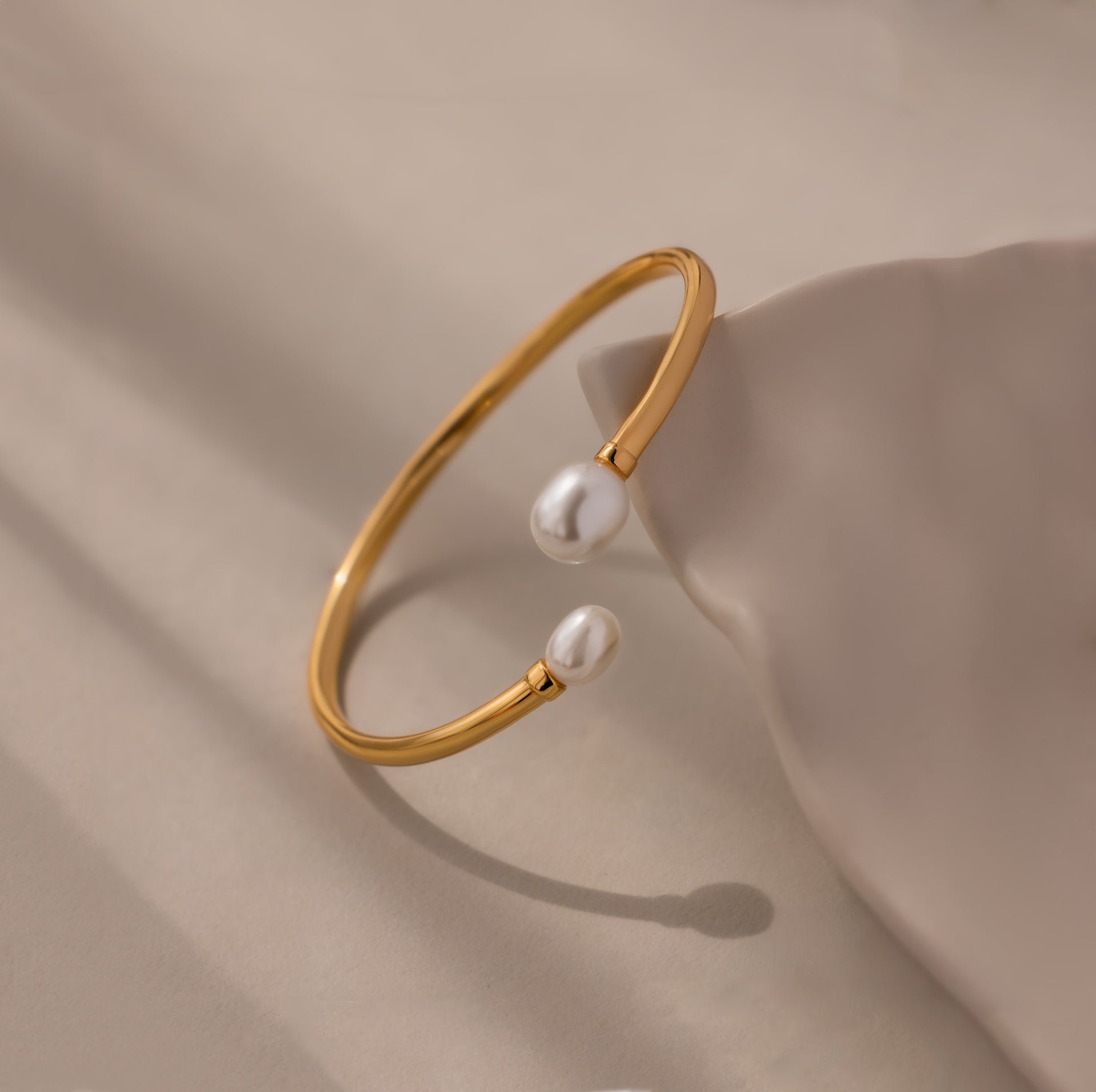 The Serenity Pearl Cuff Bracelet in 18K Gold features an open band with two white pearls on each end, elegantly displayed on a neutral beige surface—a modern statement piece.