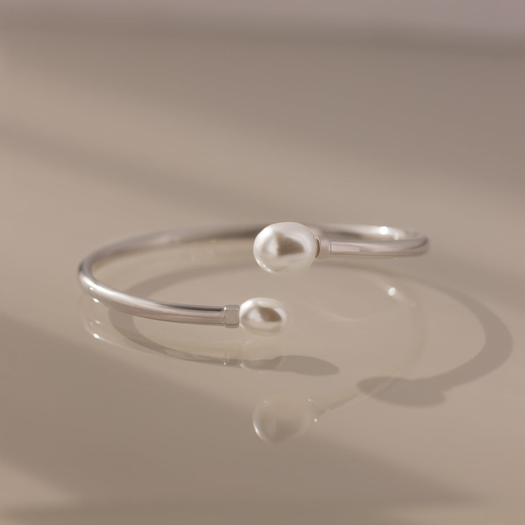 The Serenity Pearl Cuff Bracelet in Silver, featuring luminous pearls at each end, rests gracefully on a beige surface.