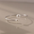 The Serenity Pearl Cuff Bracelet in Silver, featuring luminous pearls at each end, rests gracefully on a beige surface.