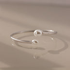 The Serenity Pearl Cuff Bracelet in Silver, featuring luminous pearls at each end, rests gracefully on a beige surface.