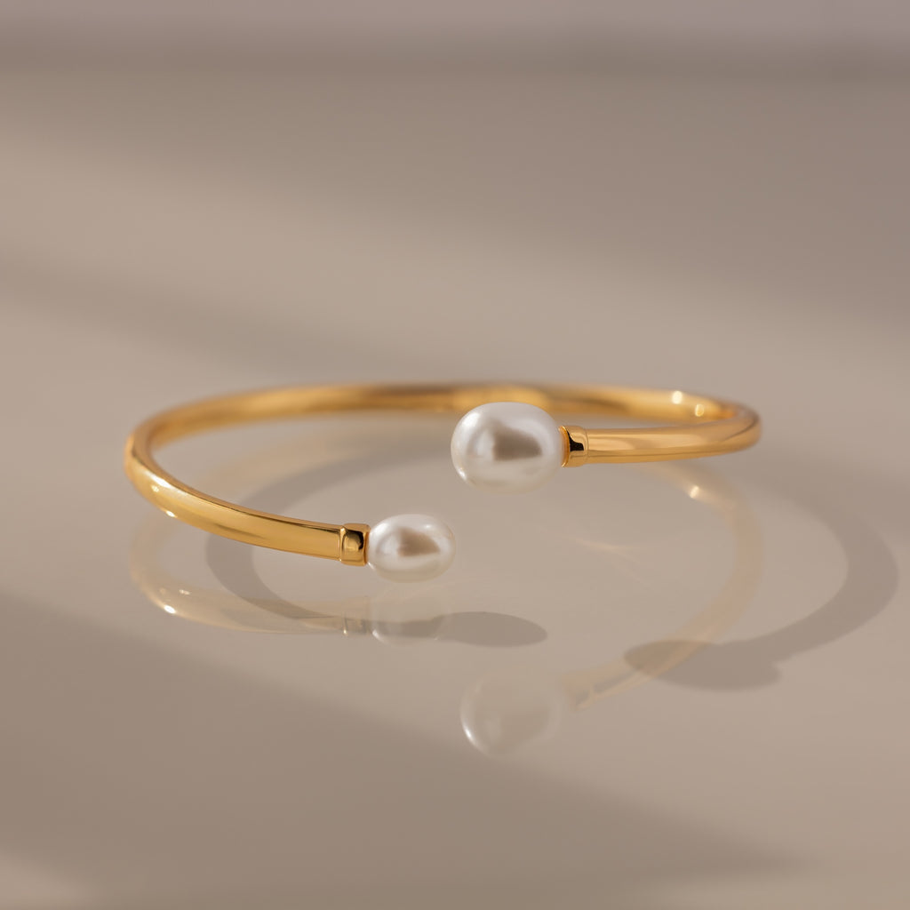 The Serenity Pearl Cuff Bracelet in 18K Gold features two white pearls on each end and a sleek open band, creating a modern and elegant jewelry piece.