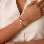 A hand wears the Serenity Pearl Cuff Bracelet—a gold open band with two pearls—resting on a cream blouse for an elegant, modern pearl jewelry look.