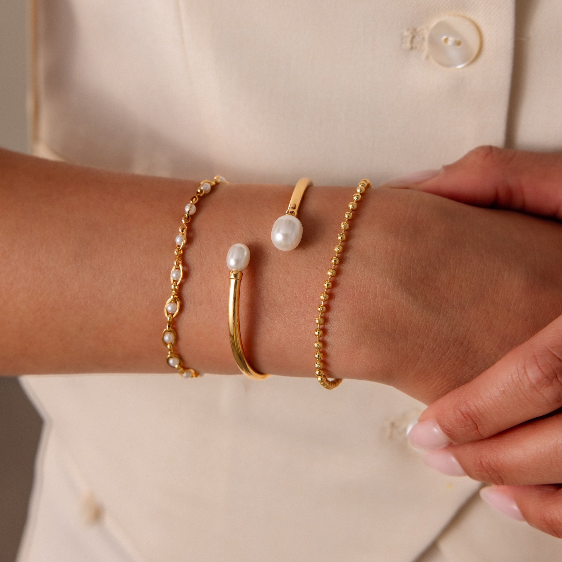 A close-up of a wrist styled with the Serenity Pearl Cuff Bracelet in 18K Gold, stacked with two gold bracelets, shown against a cream-colored outfit.