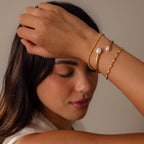Woman with closed eyes poses against a neutral background, wearing the Serenity Pearl Cuff Bracelet in 18K gold—a modern and delicate pearl jewelry piece—on her wrist.