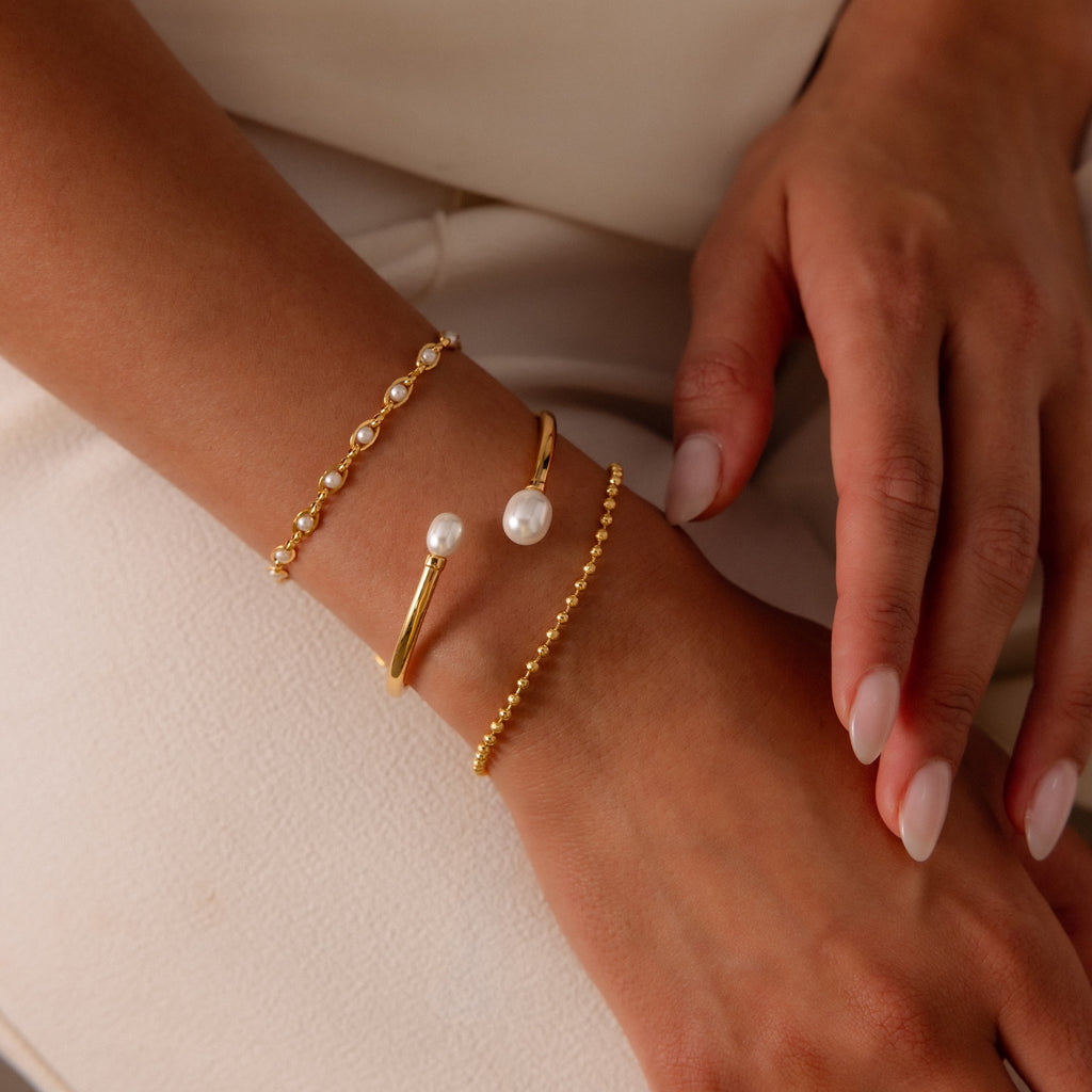A manicured hand displays the Dune Beaded Bracelet and two gold bracelets, including a layered design with pearls and polished beads, on a light fabric background.