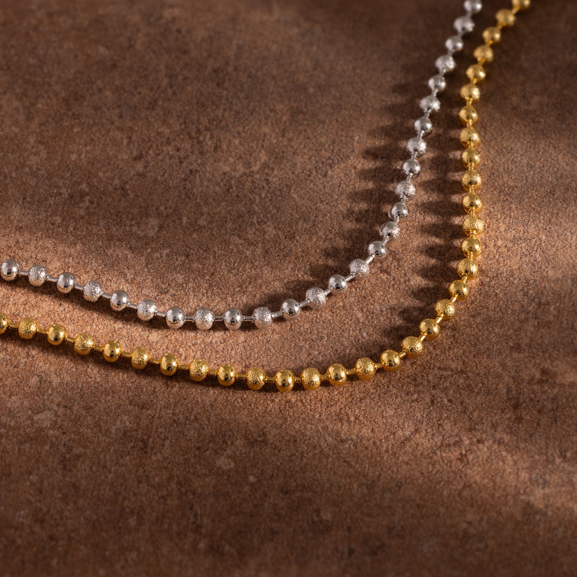 Two Dune Beaded Bracelets, with polished silver and gold beads, are displayed on a brown textured surface.