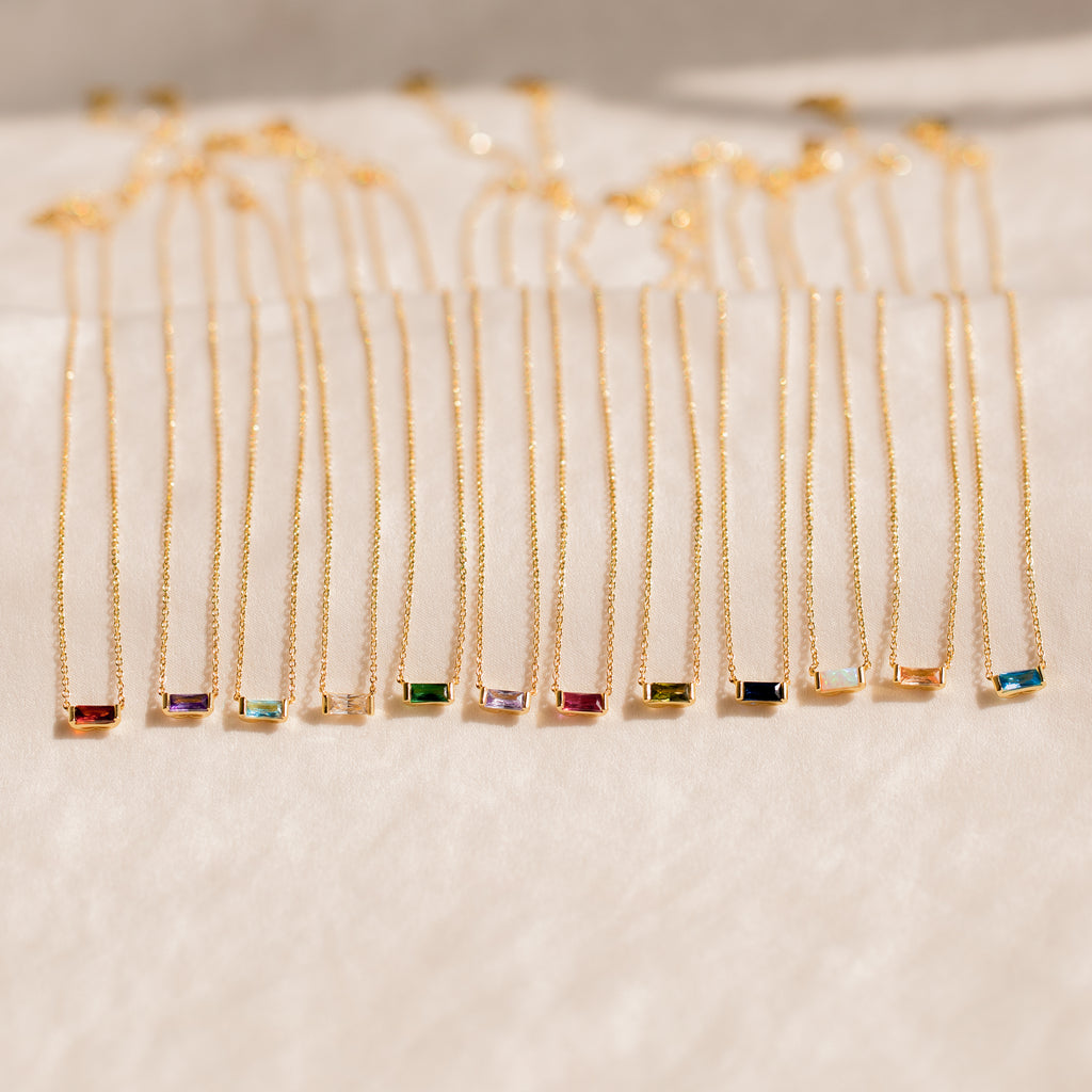 A collection of Emery Birthstone Necklaces in gold, each featuring a rectangular gemstone in a variety of vibrant birthstone colors, arranged side by side on a soft cream surface.