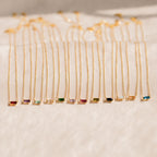 A collection of Emery Birthstone Necklaces in gold, each featuring a rectangular gemstone in a variety of vibrant birthstone colors, arranged side by side on a soft cream surface.