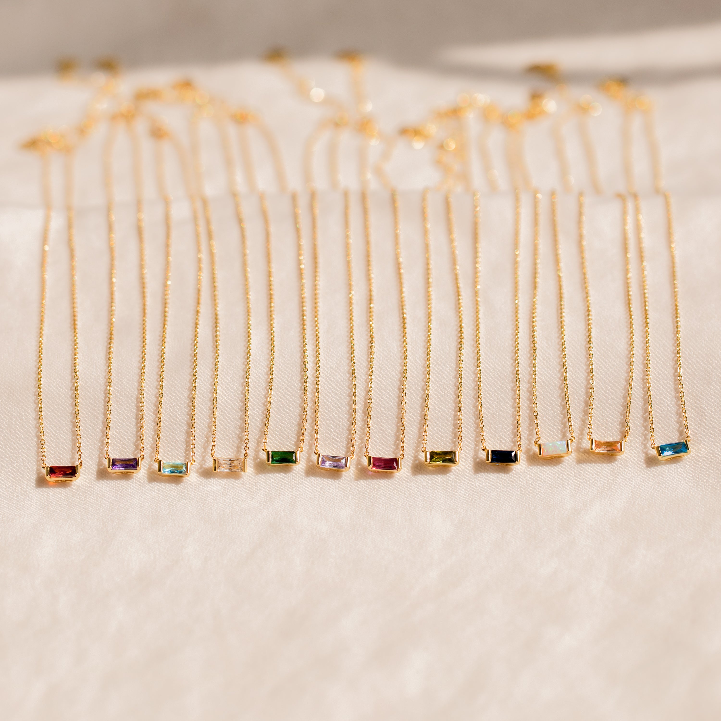 A collection of Emery Birthstone Necklaces in gold, each featuring a rectangular gemstone in a variety of vibrant birthstone colors, arranged side by side on a soft cream surface.