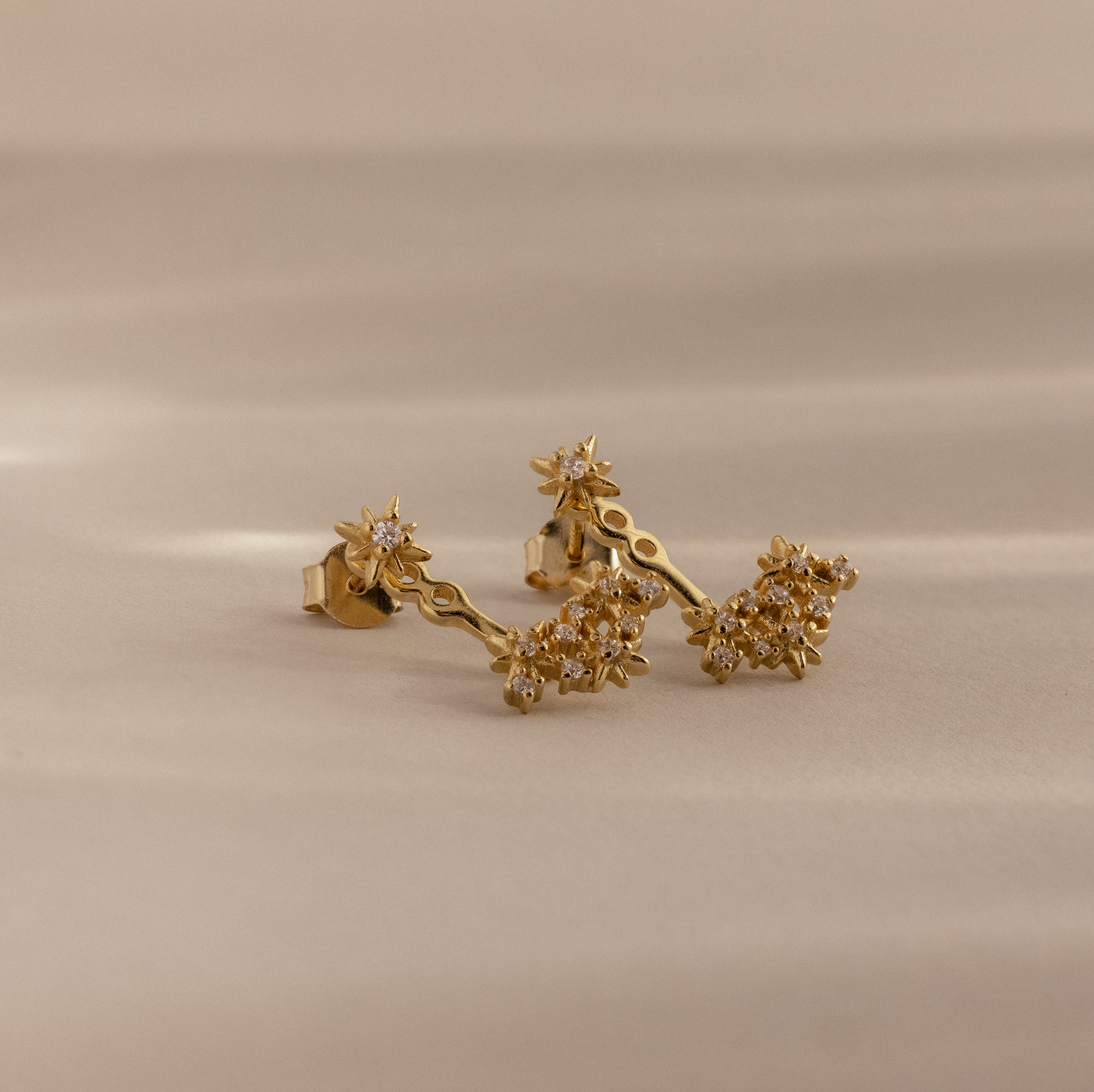 Starburst Cluster Ear Jackets in 18K Gold with small sparkling stones displayed on a soft beige surface.
