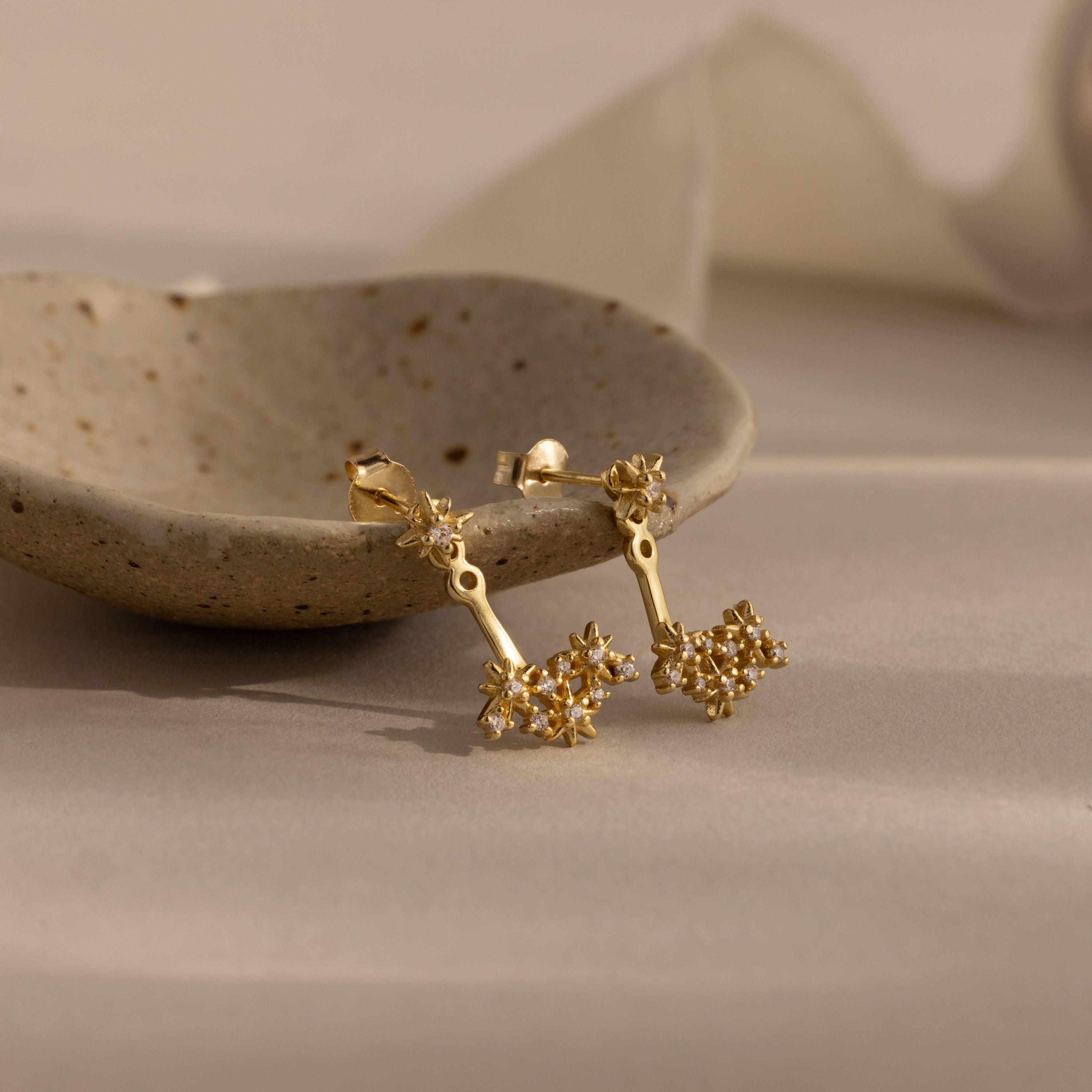 The Starburst Cluster Ear Jackets in 18K Gold evoke celestial charm with their dangling floral design, shown next to a speckled ceramic dish on a beige surface.