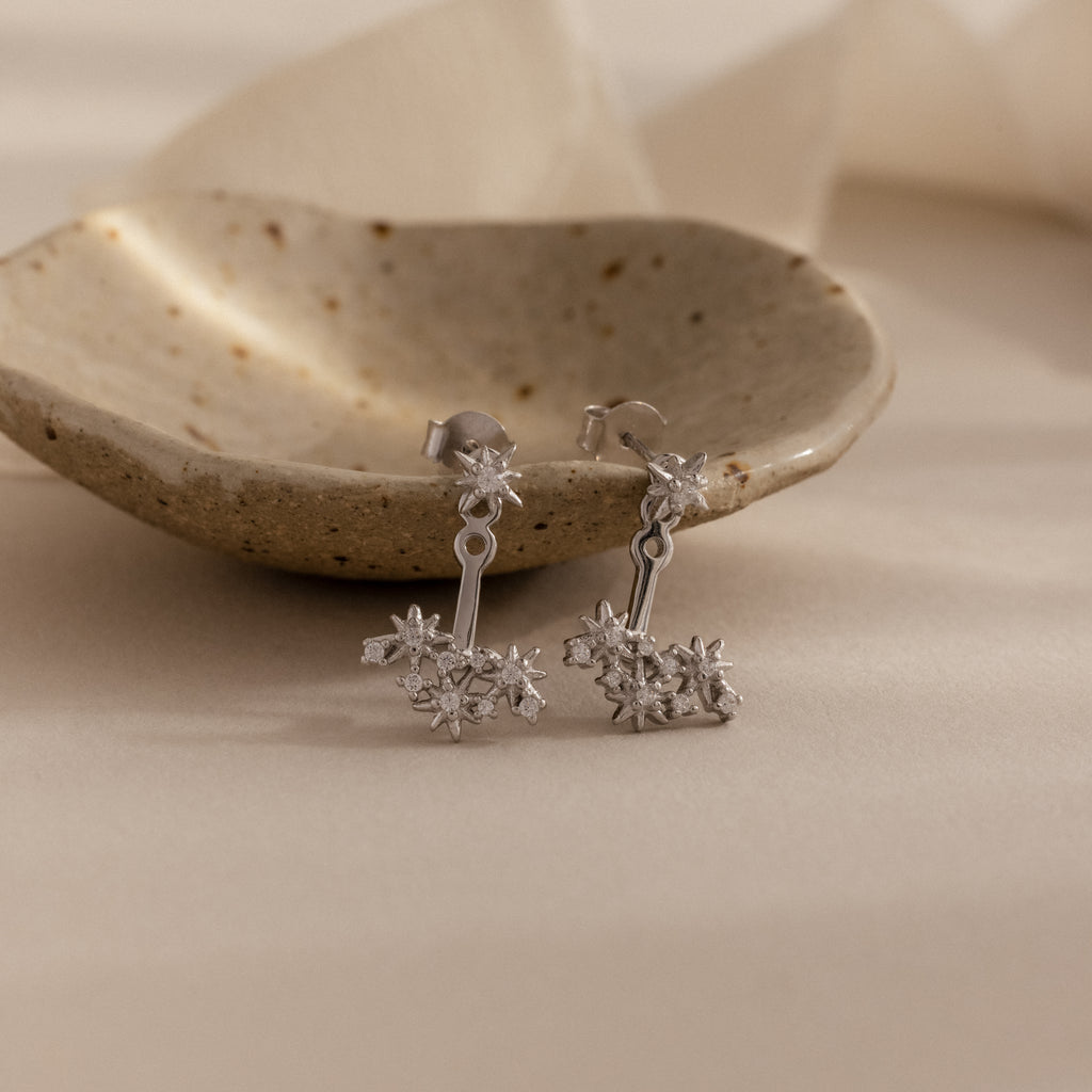 Starburst Cluster Ear Jackets in Sterling Silver, inspired by celestial motifs, displayed before a small rustic ceramic dish on a beige surface.