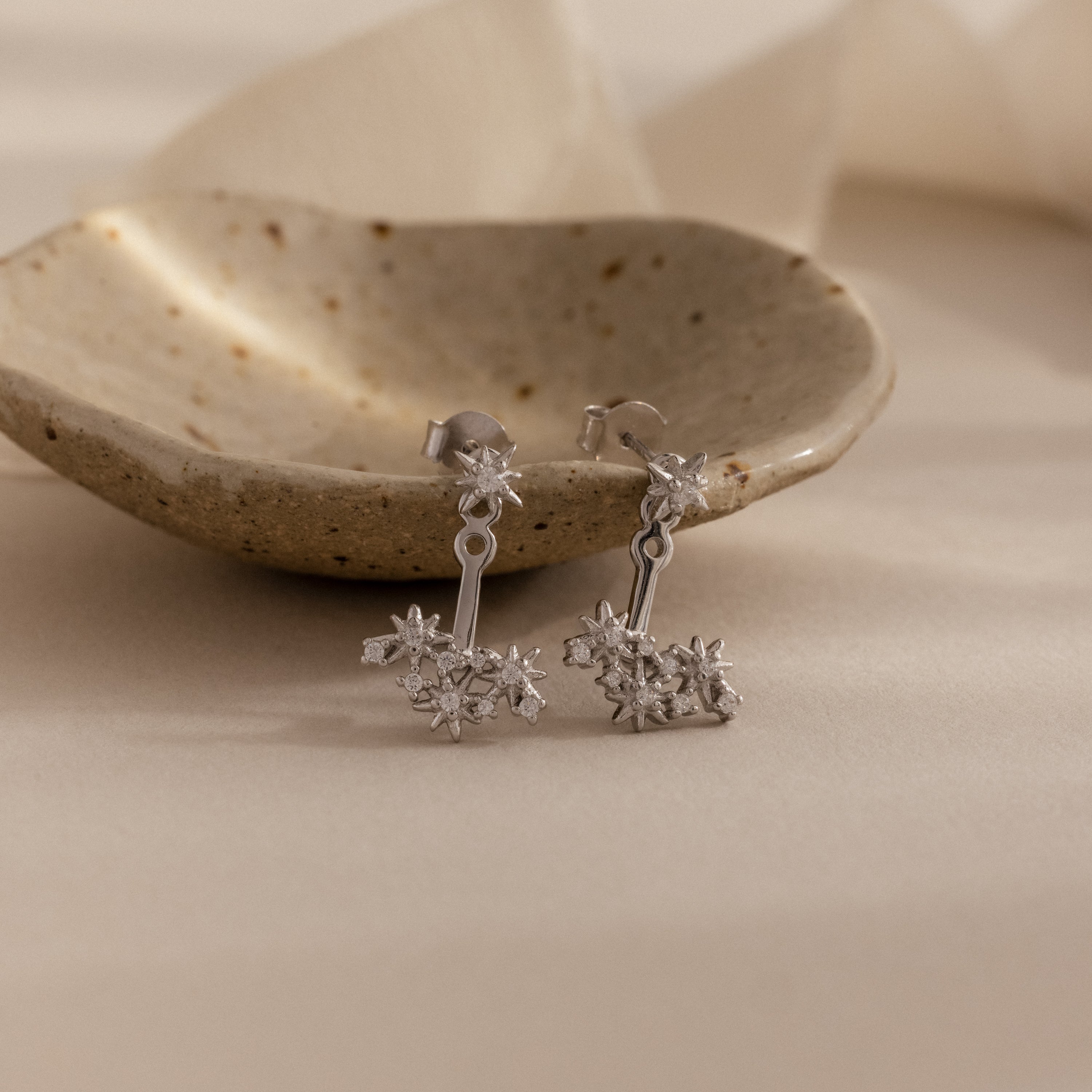 Starburst Cluster Ear Jackets in Sterling Silver, inspired by celestial motifs, displayed before a small rustic ceramic dish on a beige surface.