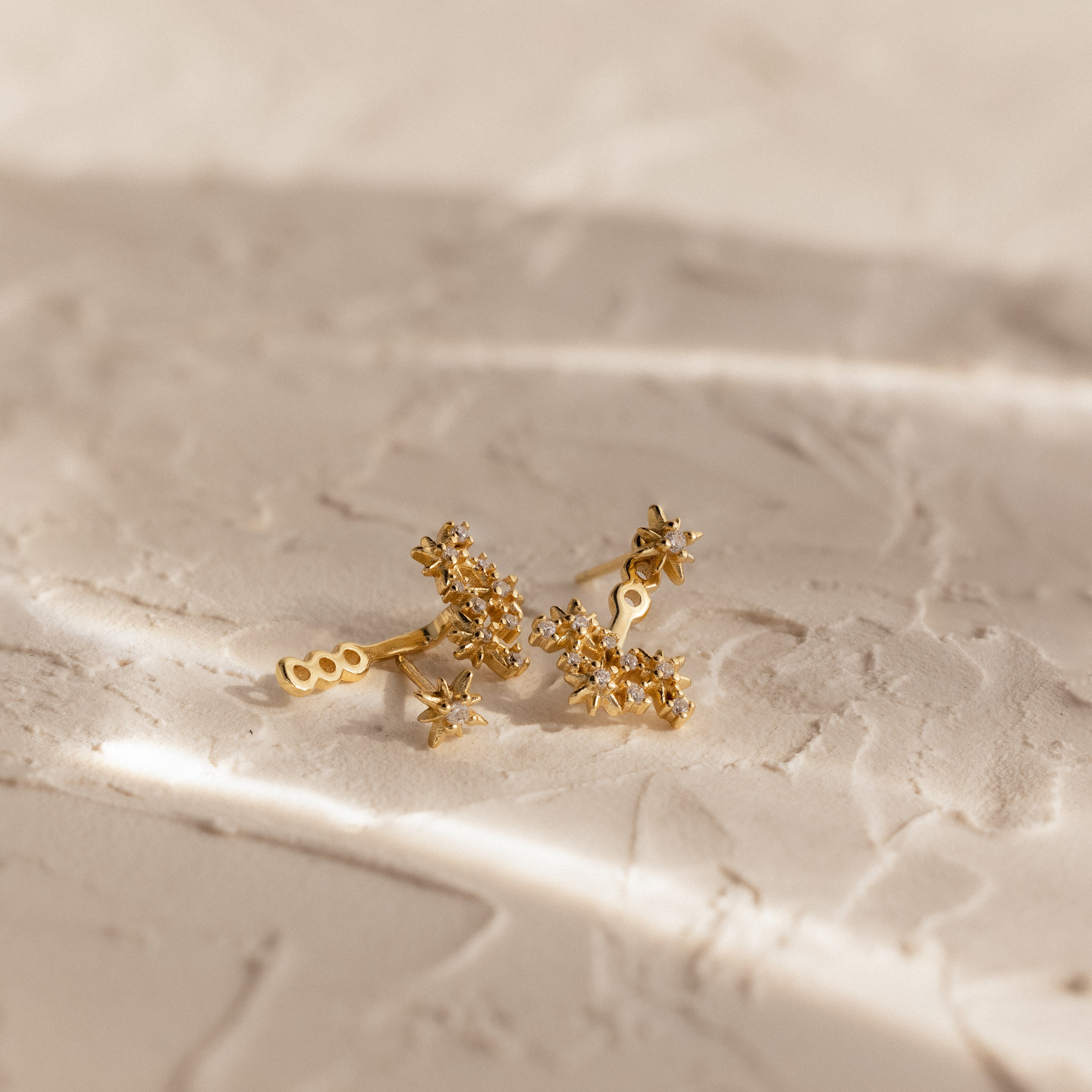 The Starburst Cluster Ear Jackets in 18K Gold, featuring a celestial star-inspired design, are displayed on a textured, light surface under soft lighting to highlight their radiant beauty.