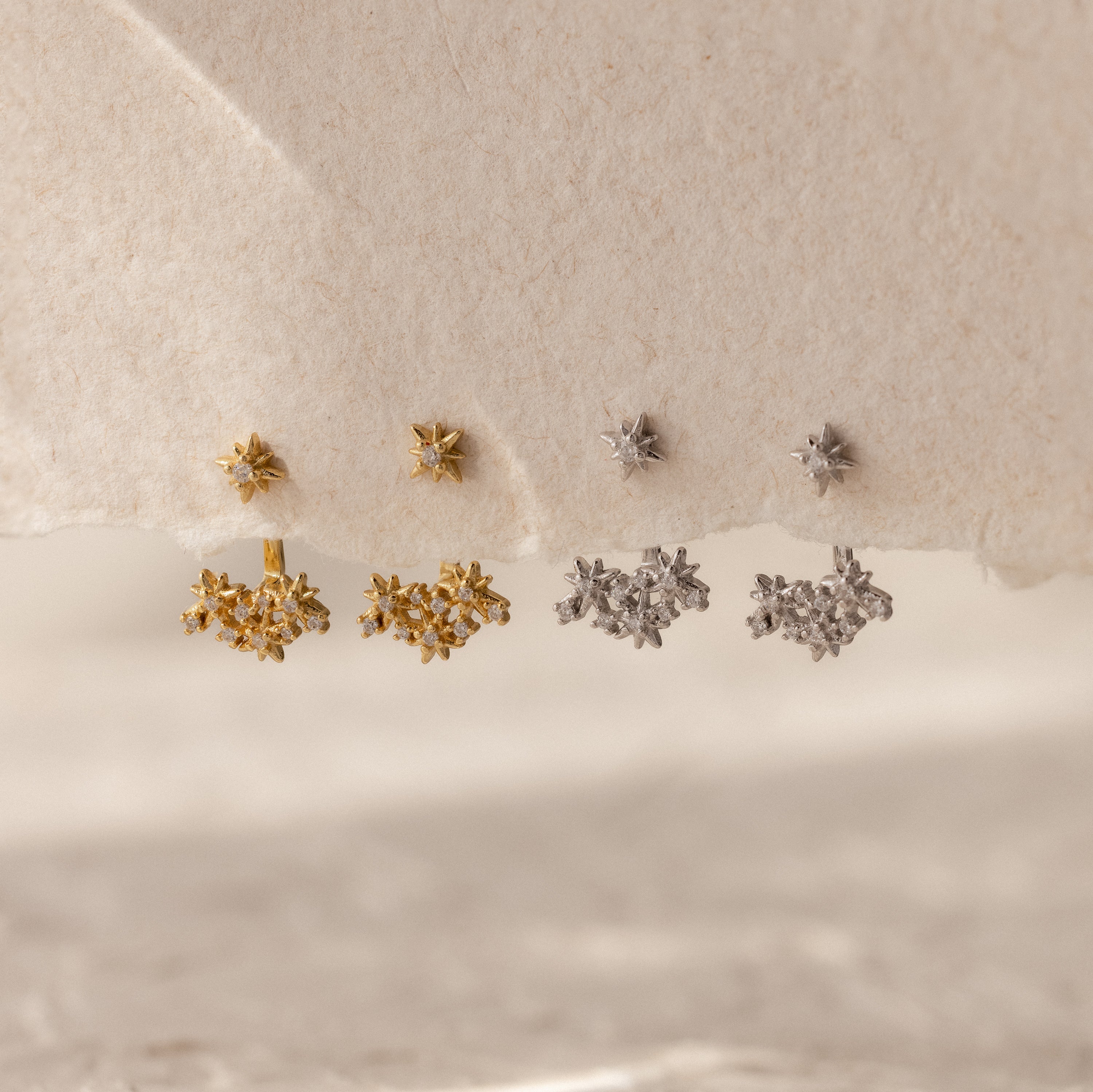 Four pairs of Starburst Cluster Ear Jackets, inspired by celestial jewelry and constellation designs, shimmer in gold and silver on a textured beige surface.