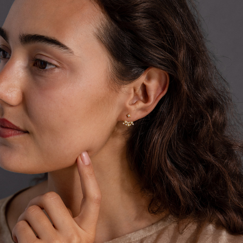 A woman with wavy brown hair wears celestial jewelry—star-shaped earrings inspired by Starburst Cluster Ear Jackets in 18K Gold—as she gently touches her chin.