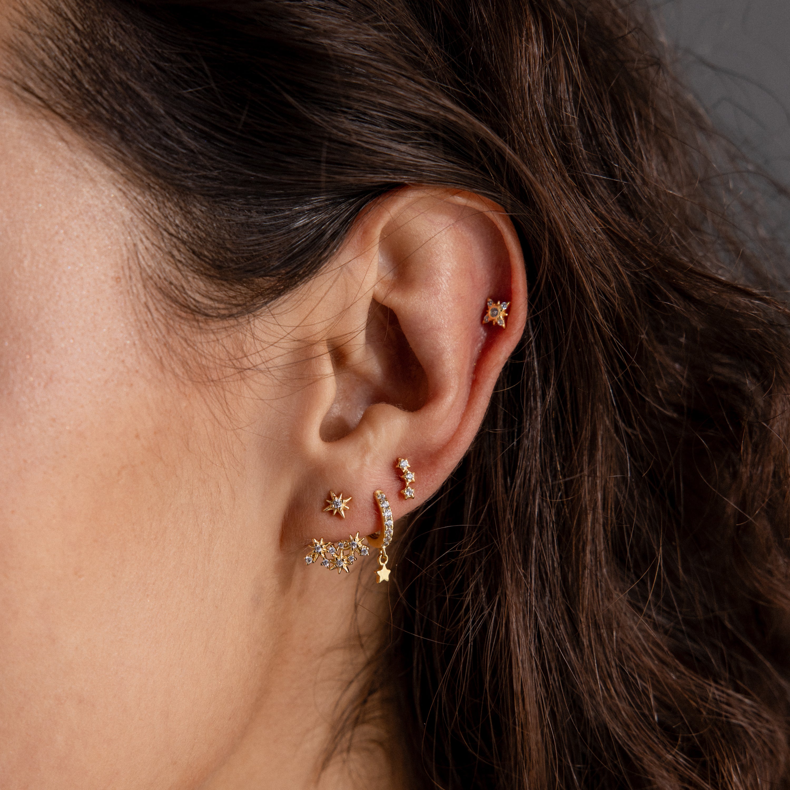 Close-up of a woman's ear adorned with celestial jewelry—multiple gold constellation earrings, including the Starburst Cluster Ear Jackets in 18K Gold and other pieces with stars and small gemstones.